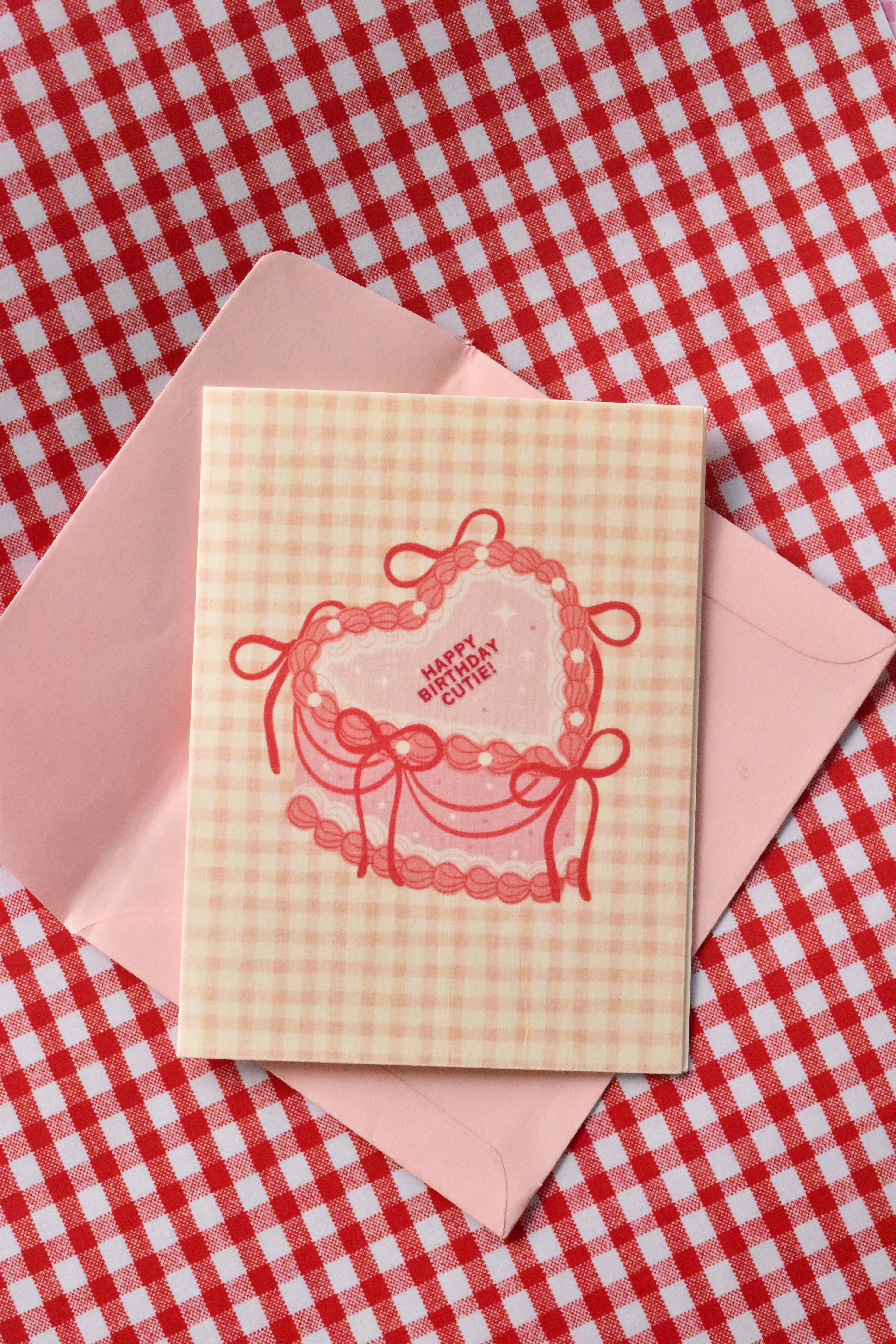  A top-down shot of a square greeting card that reads "Happy Birthday Cutie!" on a pink, heart-shaped layer cake illustration. The card has a light pink and cream gingham background and sits inside a matching light pink envelope. 