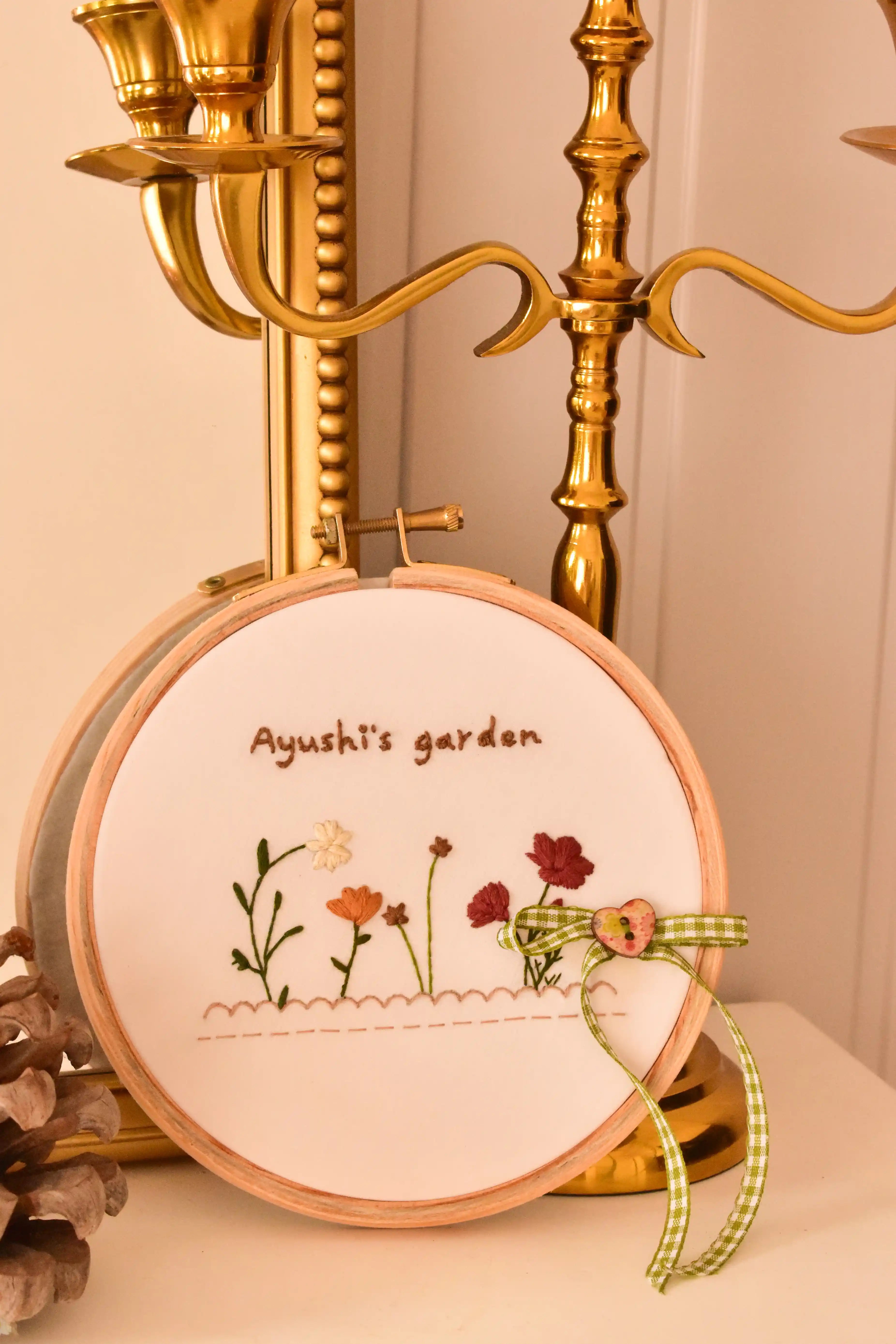  A custom hand embroidered 6-inch hoop reading "Ayushi's garden" with flowers, displayed next to a golden candelabra.