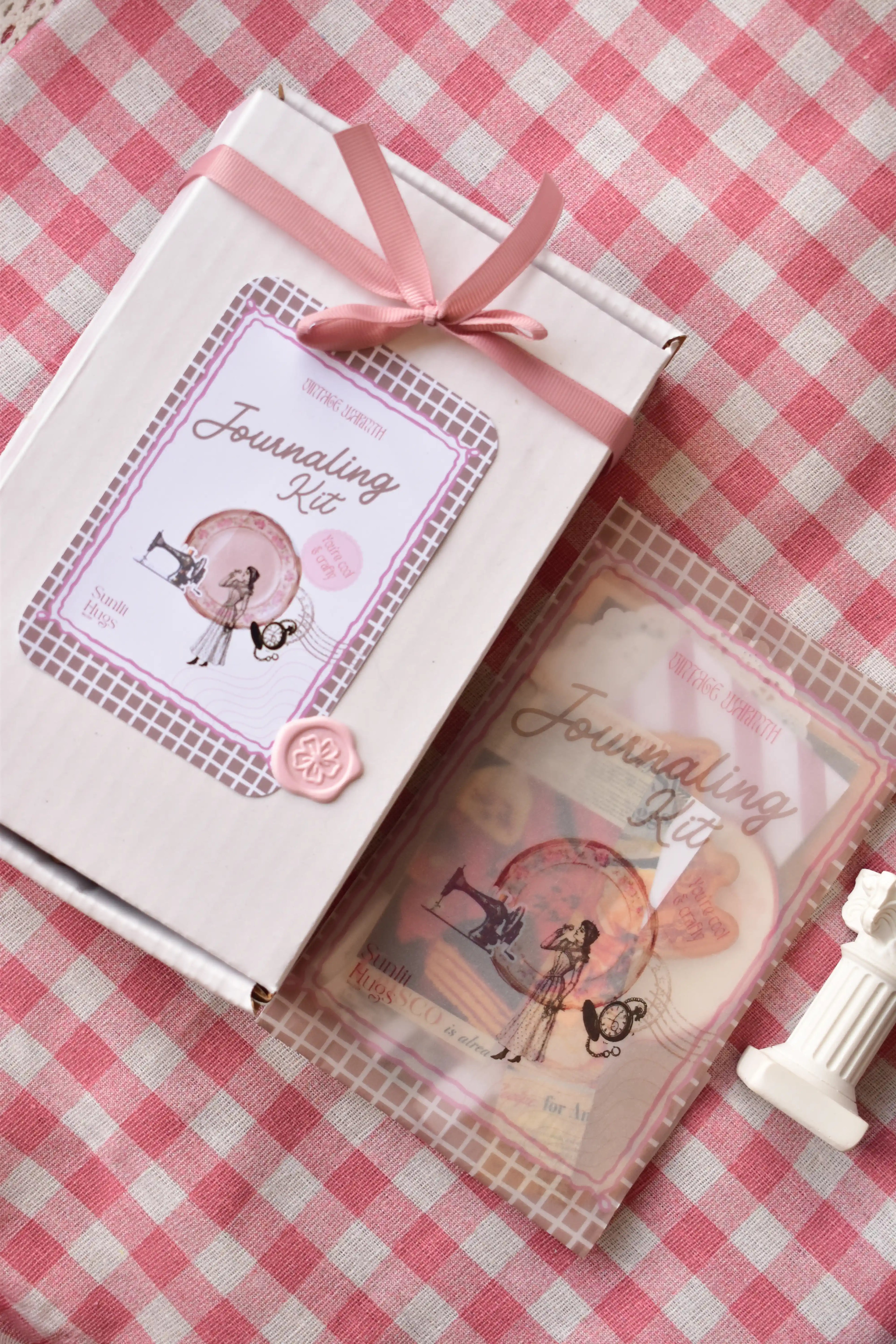  A white Journaling Kit box tied with a pink ribbon, next to a sample of the kit's vintage ephemera in a clear package.