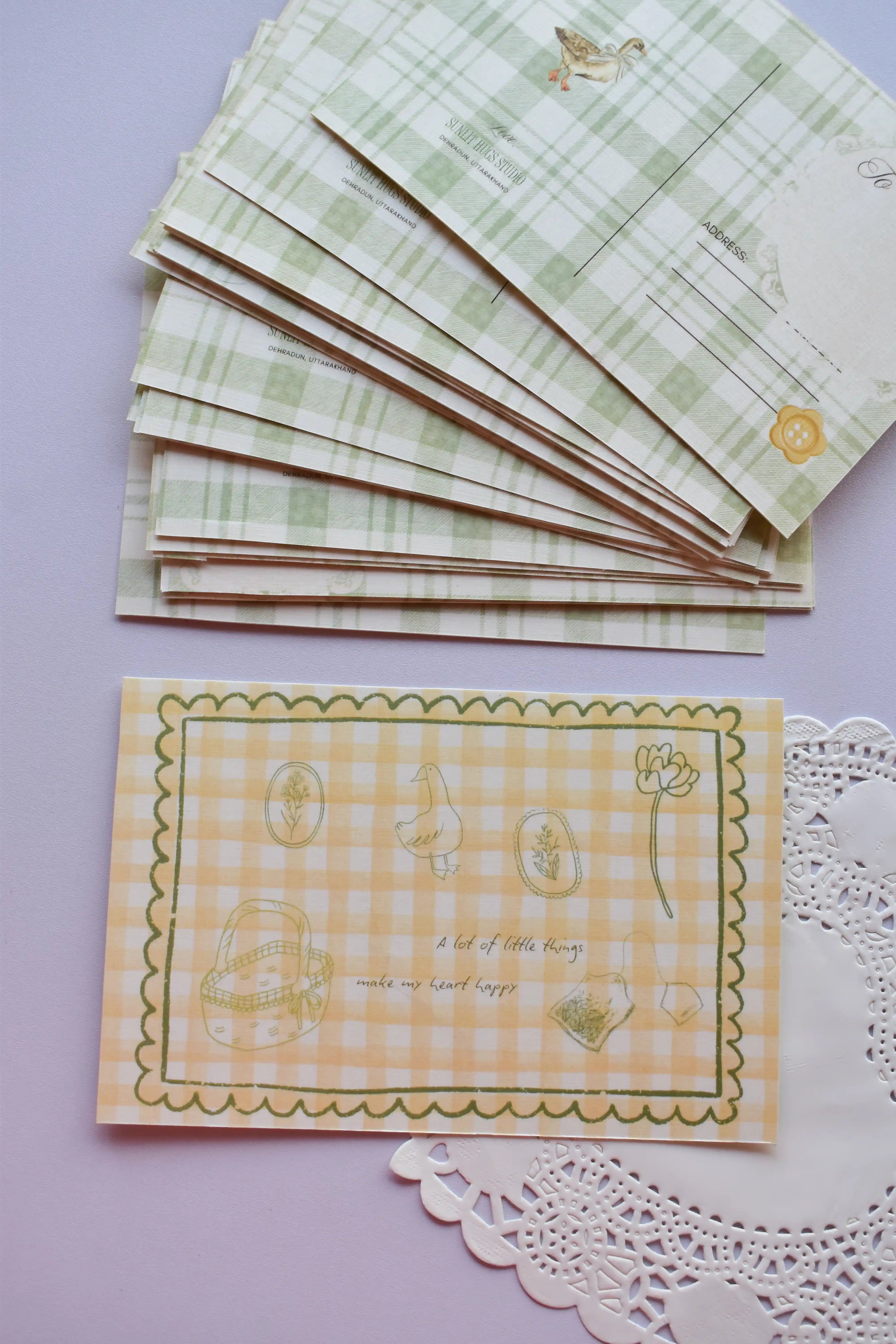 Close-up of the front of the postcard featuring the yellow and white wide gingham background with the line-art sketches of a basket, duck, and flowers, and the quote, "A lot of little things make my heart happy."