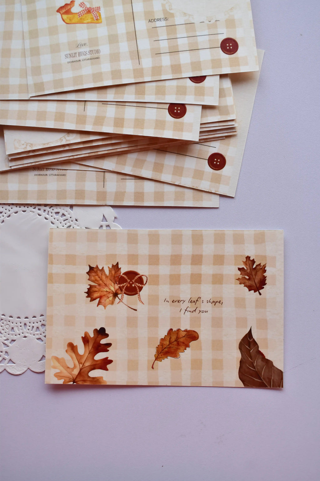 A stack of postcards showing the address and pie slice illustration on the top few cards, with one card laid flat displaying the front design of brown autumn leaves, an acorn, and the quote "In every leaf's shape, I find you."