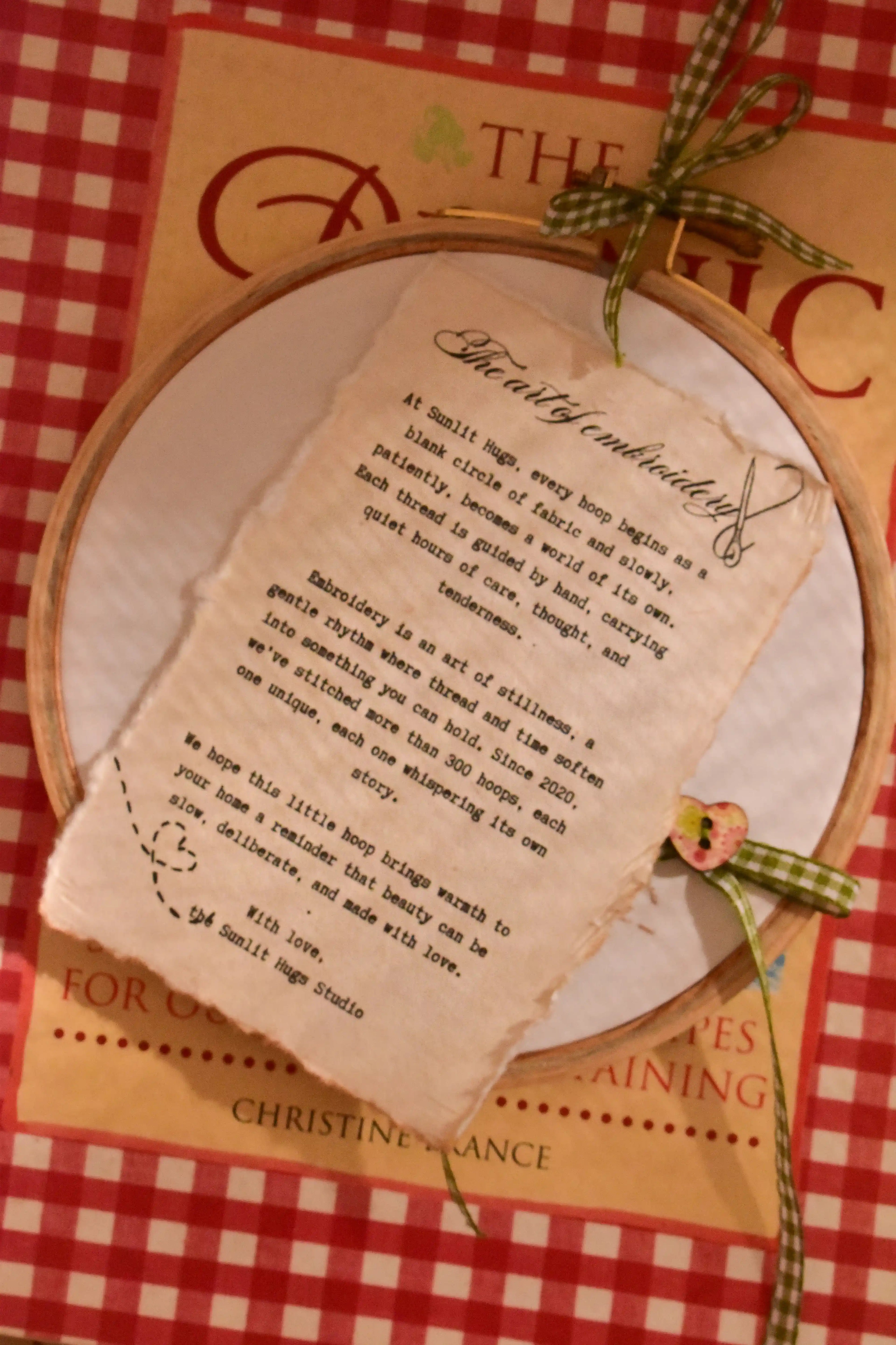 Hand embroidered hoop featuring a handwritten note about "The art of embroidery," resting on a red gingham book cover.