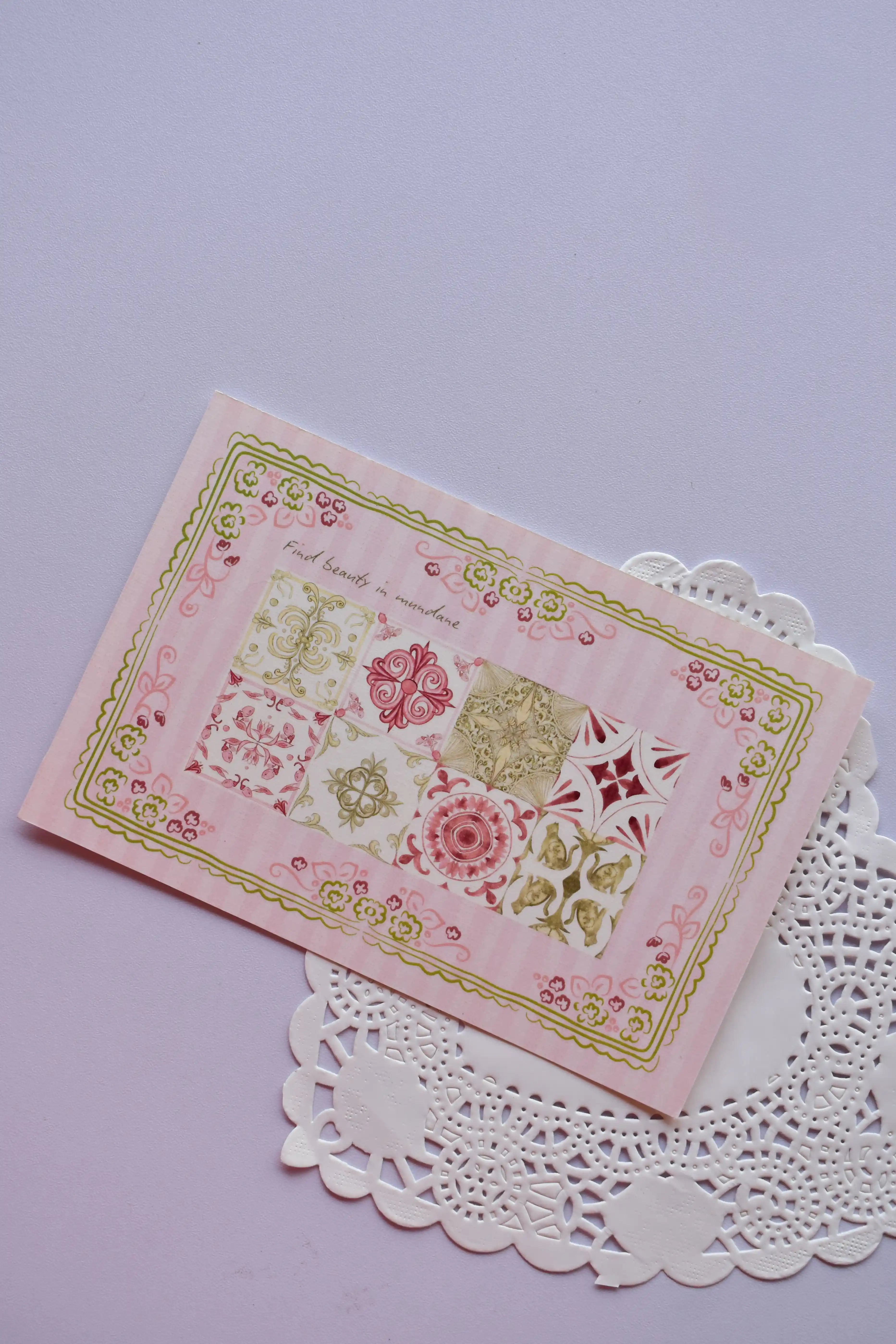 Close-up of a horizontal postcard featuring a pink background with nine square vintage tile patterns in red, gold, pink, and white. The entire mosaic is framed by a decorative green floral border, with the text "Find beauty in mundane." The card rests on a white paper doily.