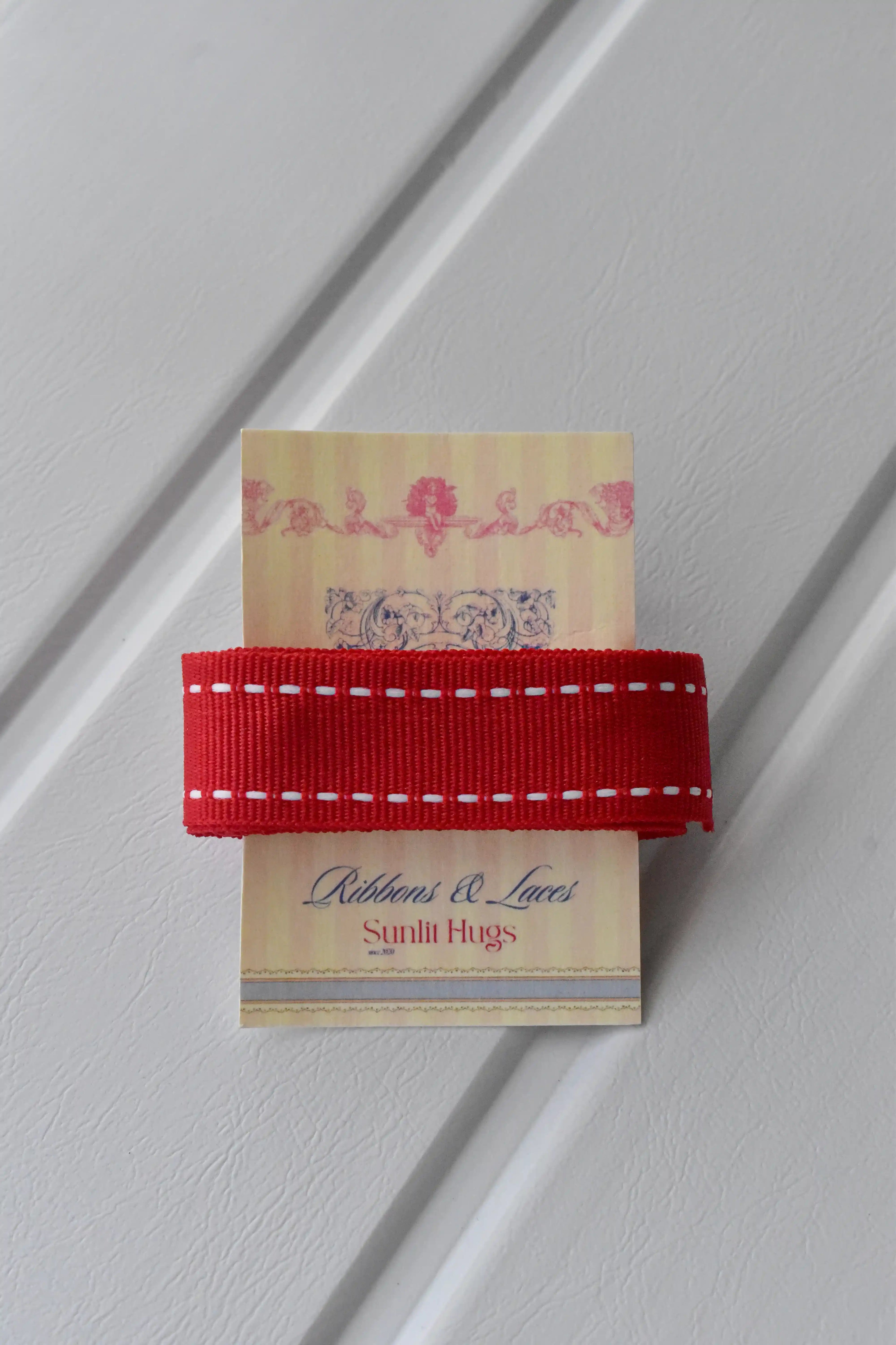 This image shows the texture and detail of the Berry Stitch ribbon, highlighting the red grosgrain material and the white stitching along the edges, with the "Sunlit Hugs Ribbons & Laces" tag as a brand identifier.