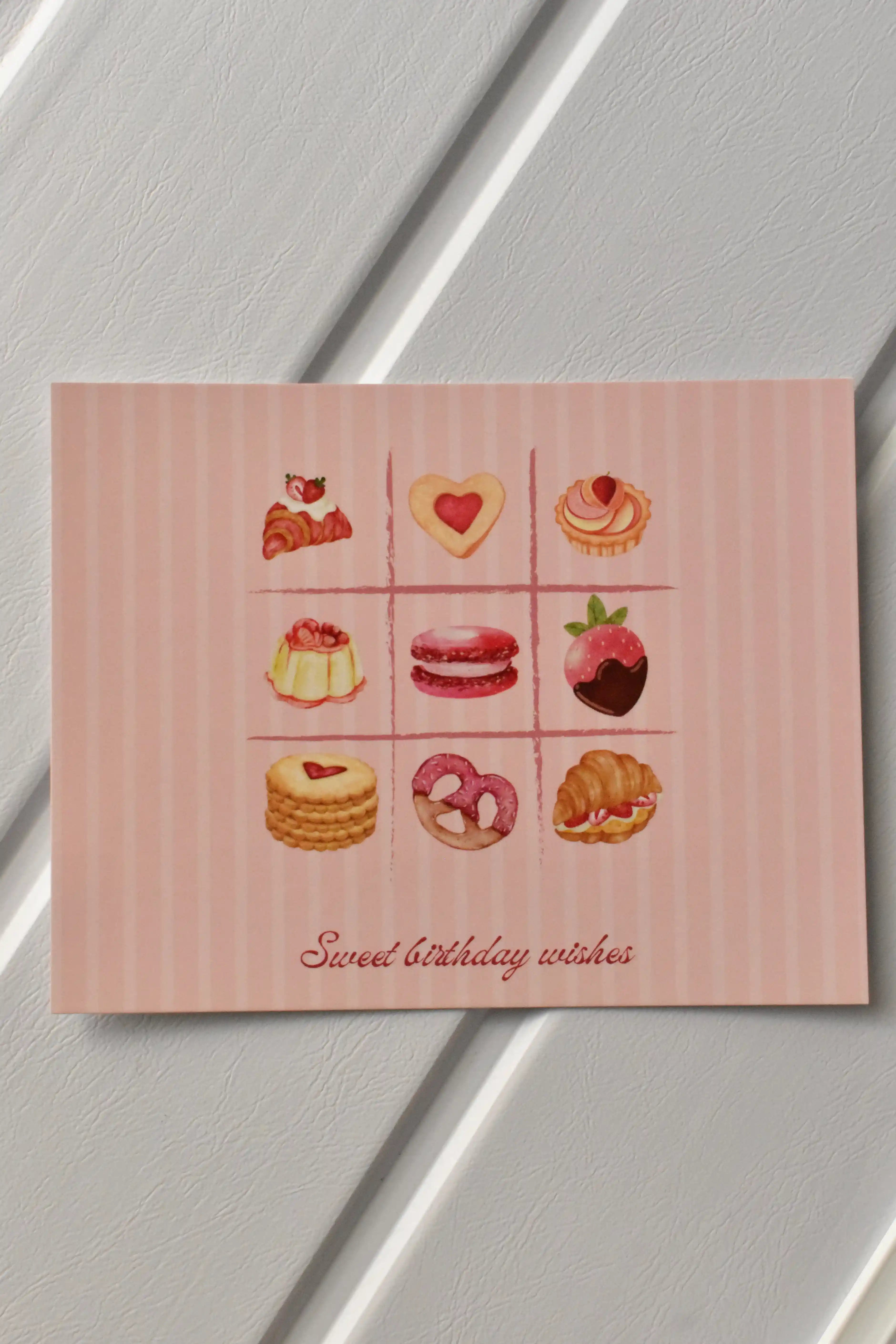 This image showcases the main front design of the Birthday Bakes Postcard, highlighting the vibrant grid of nine whimsical desserts and the soft pink striped background.