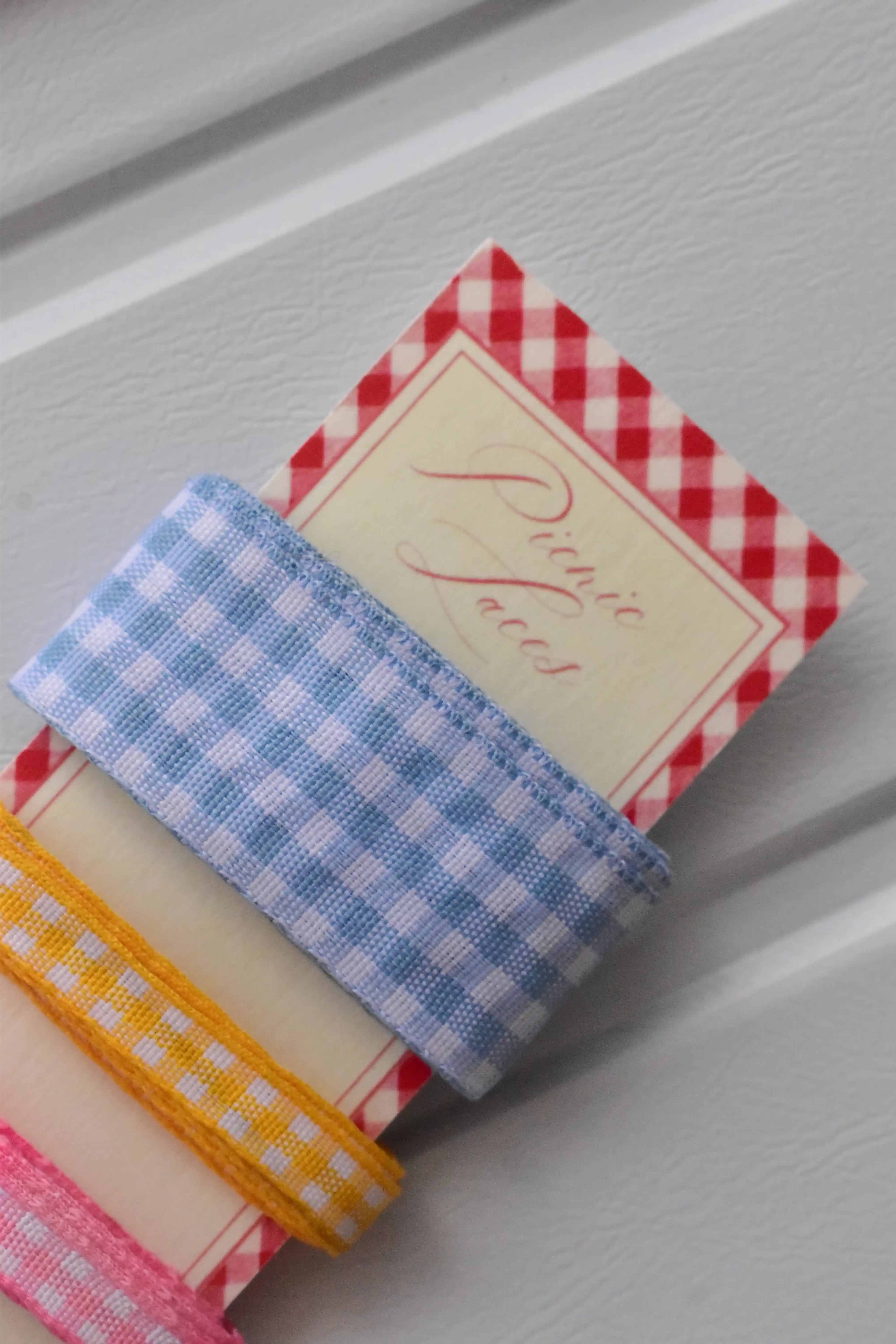 Angled close-up showing the blue, yellow, and pink narrow gingham ribbons wrapped around the top corner of the "Picnic Laces" display card.