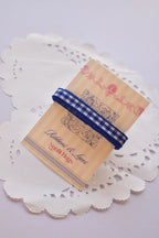 Narrow navy blue and white gingham ribbon wrapped tightly around a "Sunlit Hugs" card, resting on a decorative white lace doily.