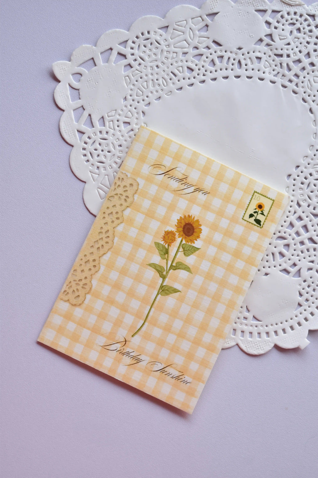 Close-up of a small, cheerful greeting card featuring a yellow and white gingham check pattern. The center has a small square with three miniature sunflowers, and the text "Sunlit Hugs" is at the bottom left. The card is positioned on a white paper doily.