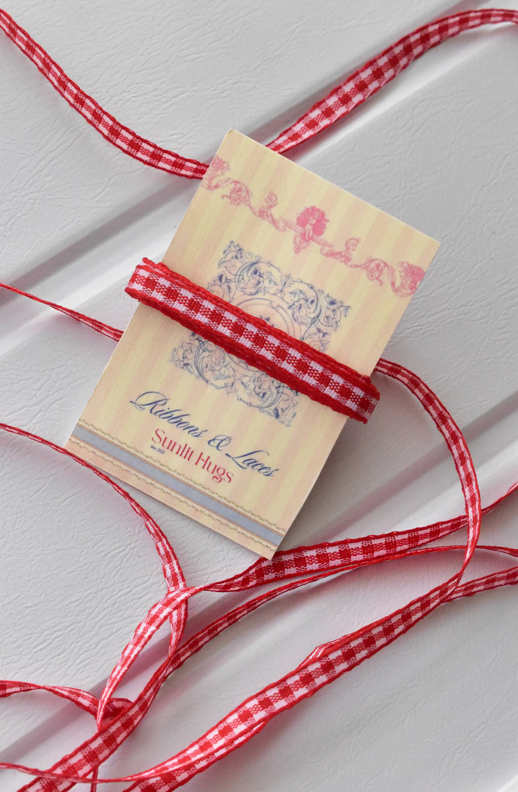 This image features the Cherry Picnic narrow ribbon with a long, flowing strand unravelled, showcasing its full red and white checkered pattern and texture. A small section is also wrapped around a "Sunlit Hugs" brand tag.
