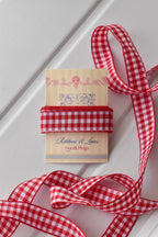 This close-up highlights the texture and design of the Cherry Picnic Wide ribbon. The image shows the ribbon swatch on the "Sunlit Hugs" brand tag with a long piece unraveled, giving a full view of its wide red and white checkered pattern.