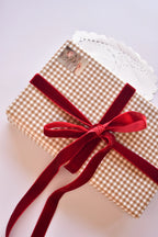 A gift box wrapped in tan checkered paper tied with a generous cherry red velvet ribbon bow.