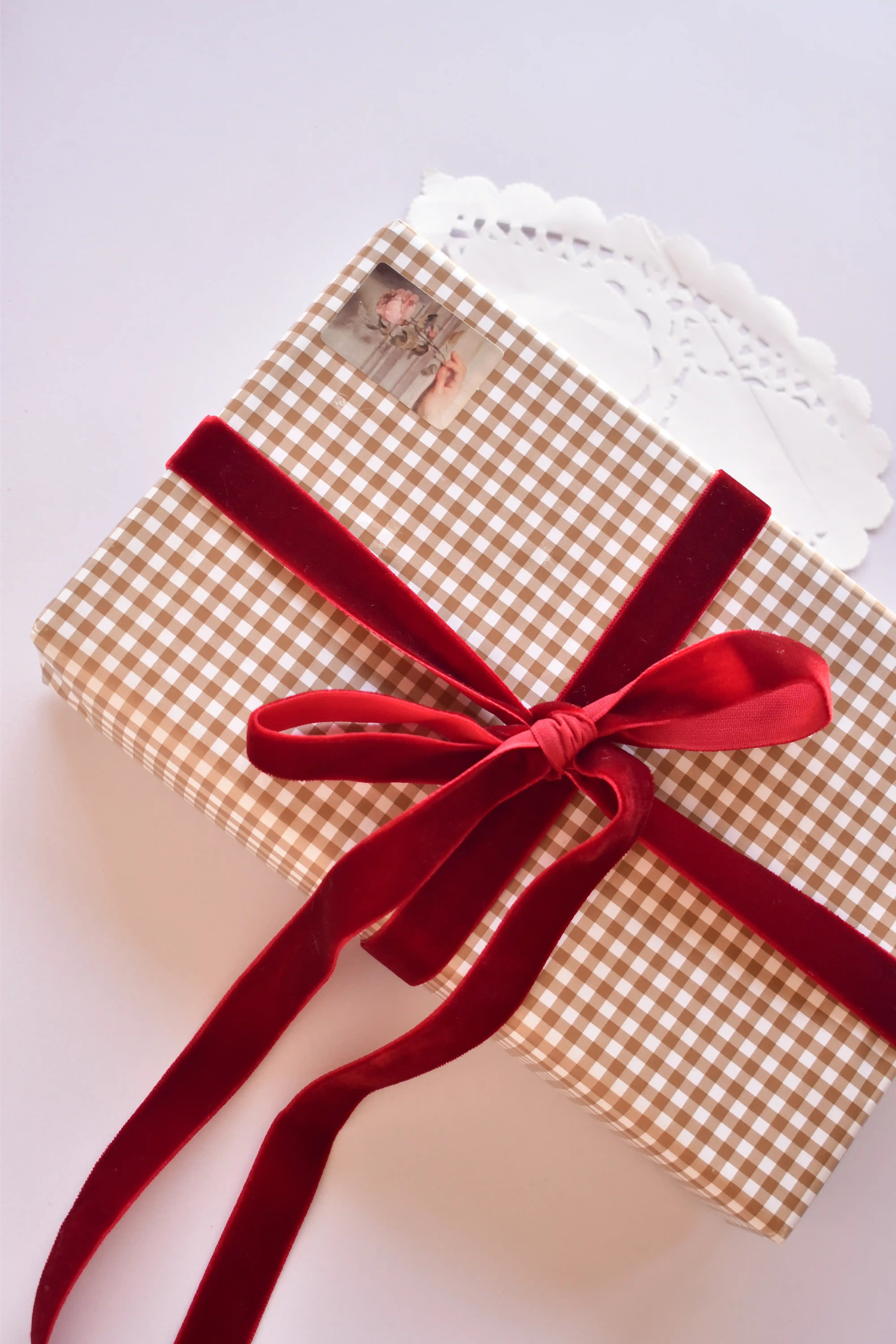 A gift box wrapped in tan checkered paper tied with a generous cherry red velvet ribbon bow.