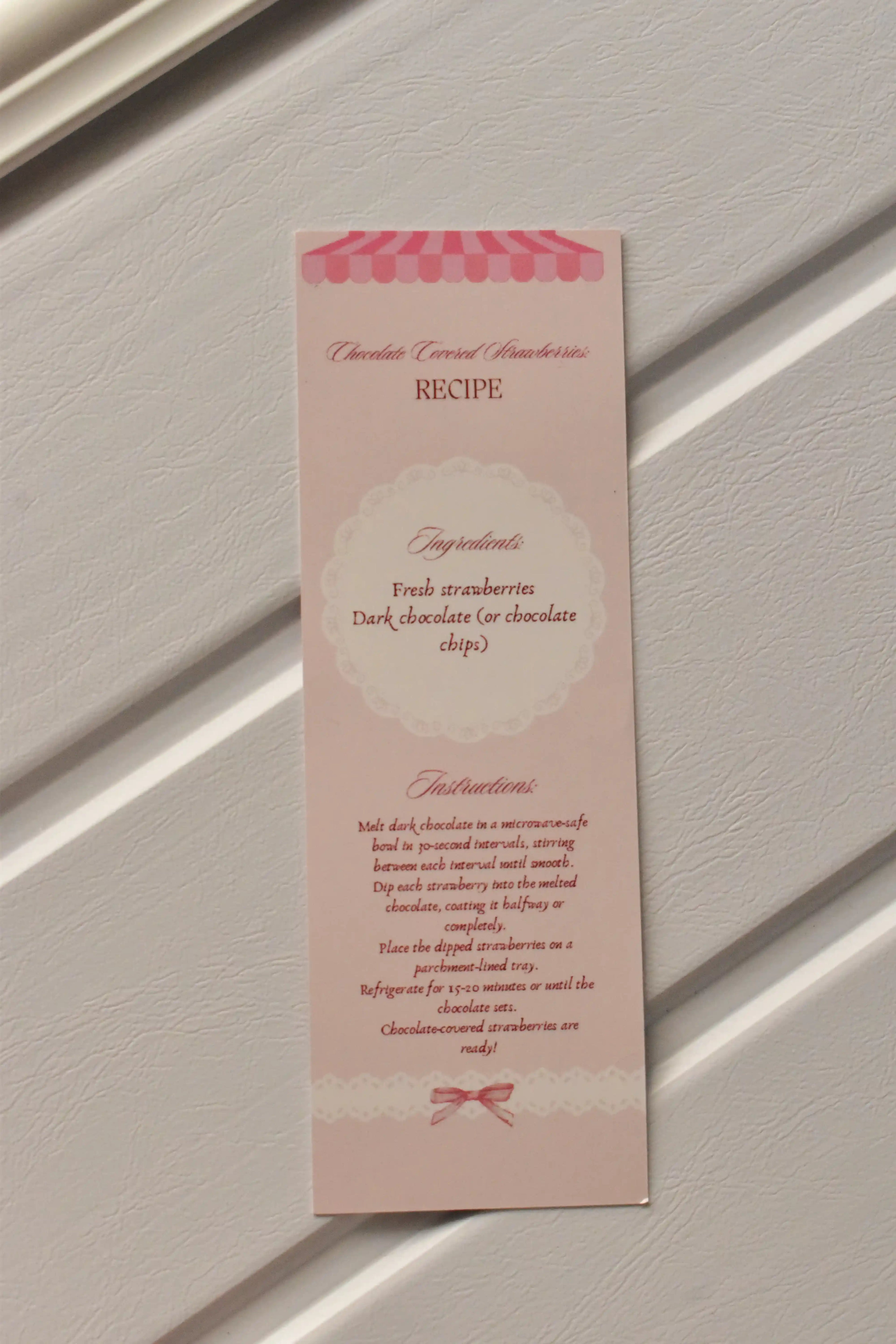 This image focuses on the practical element, displaying the detailed Chocolate Covered Strawberries Recipe printed on the back of the pastel pink bookmark, ready for kitchen inspiration.