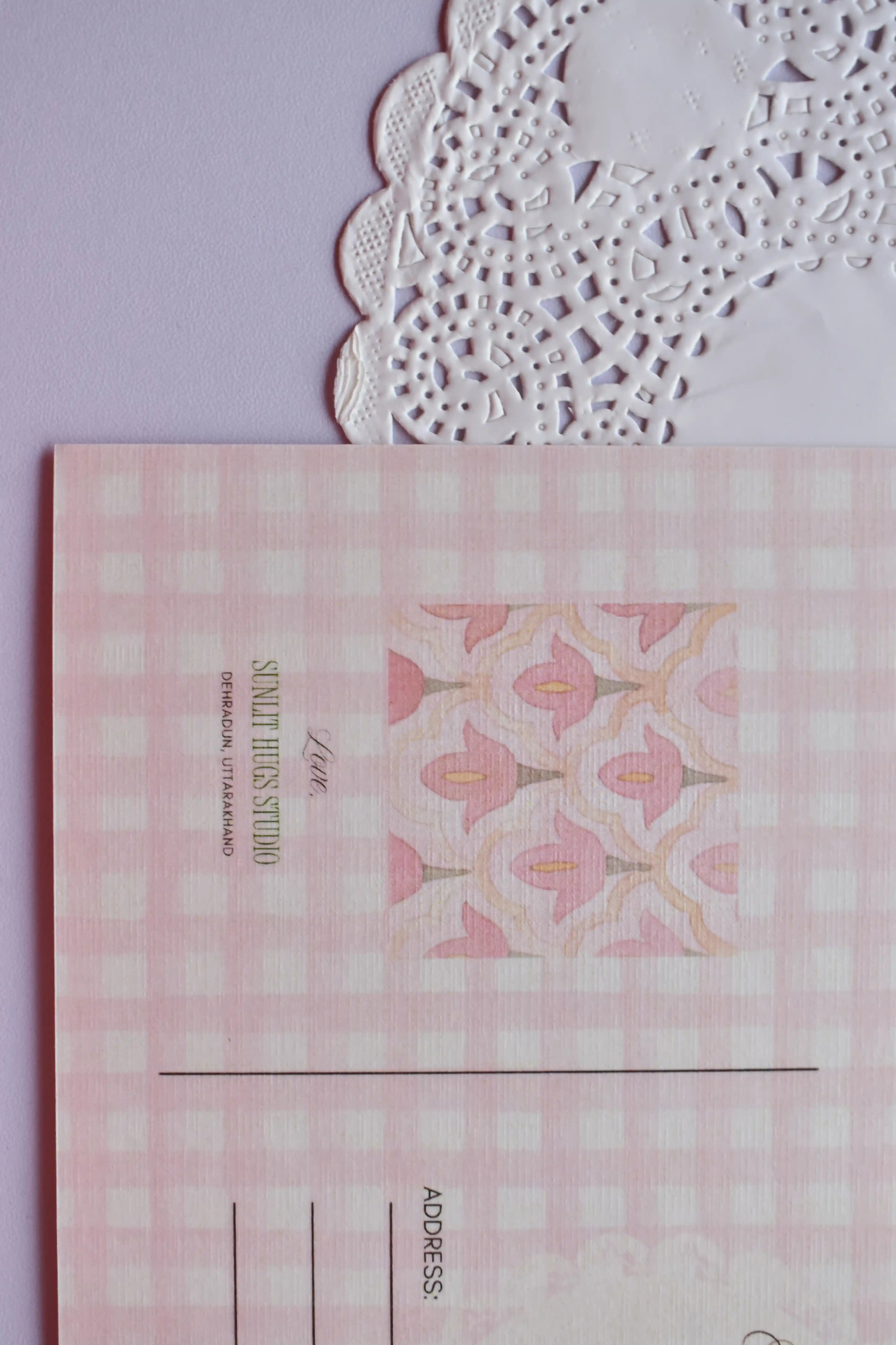 Extreme close-up of the postcard reverse, showing the pink and white wide gingham pattern and the central pink floral bell pattern square. The text "SUNLIT HUGS STUDIO" is visible.