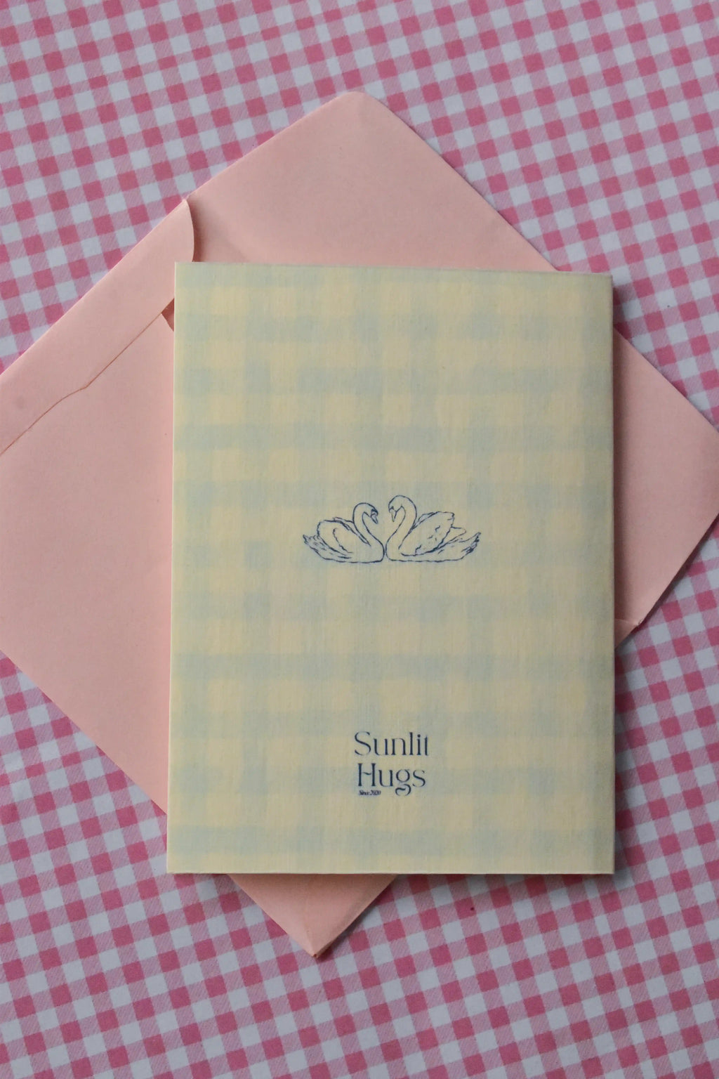 This image displays the back of the card, featuring a charming blue illustration of two swans that symbolise enduring connection, along with the Sunlit Hugs logo, sitting on a pink envelope.
