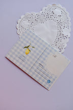 The back of a postcard with a light blue and white wide gingham pattern, showcasing a single hanging lemon illustration near the address lines, set against a heart-shaped doily.