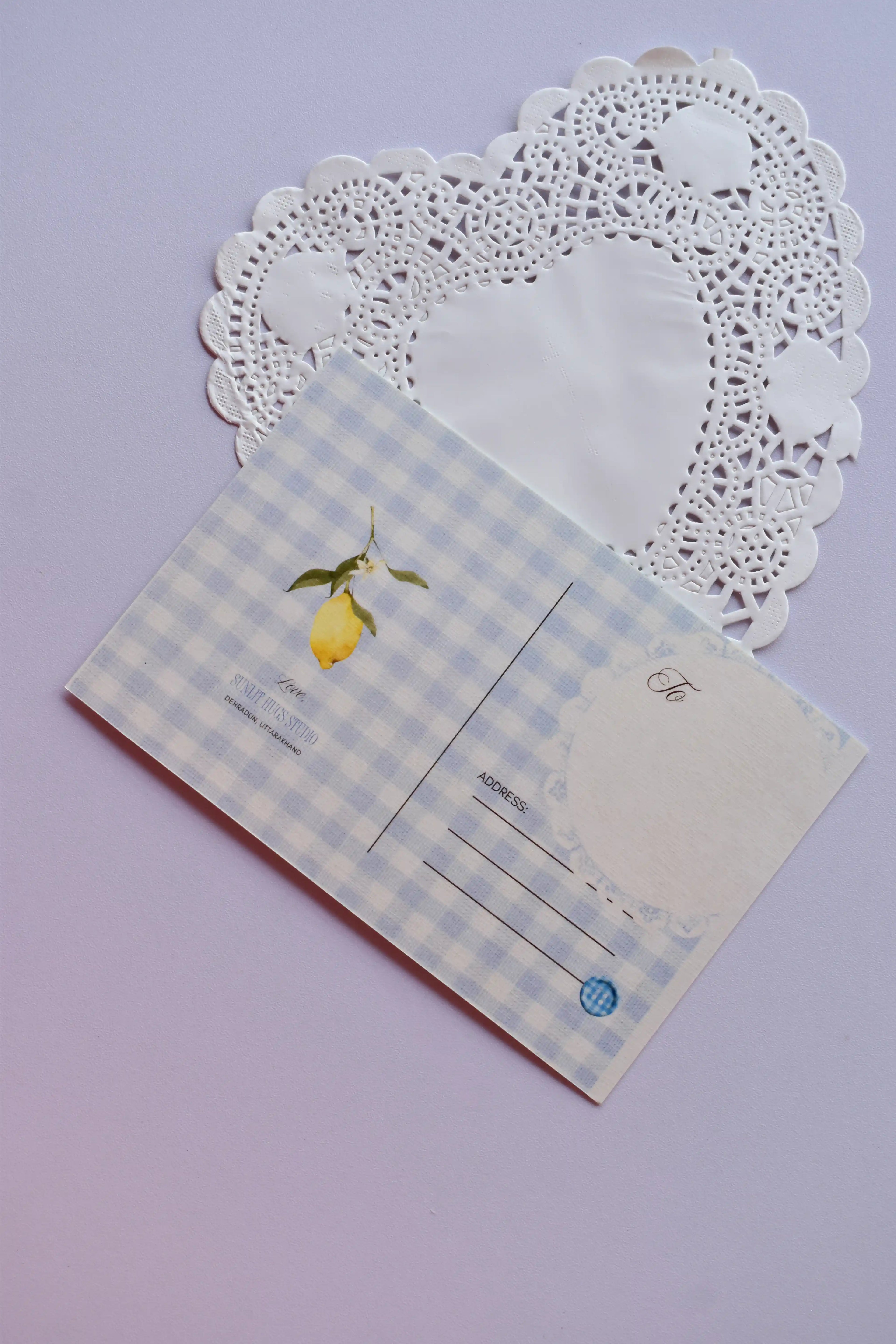 The back of a postcard with a light blue and white wide gingham pattern, showcasing a single hanging lemon illustration near the address lines, set against a heart-shaped doily.