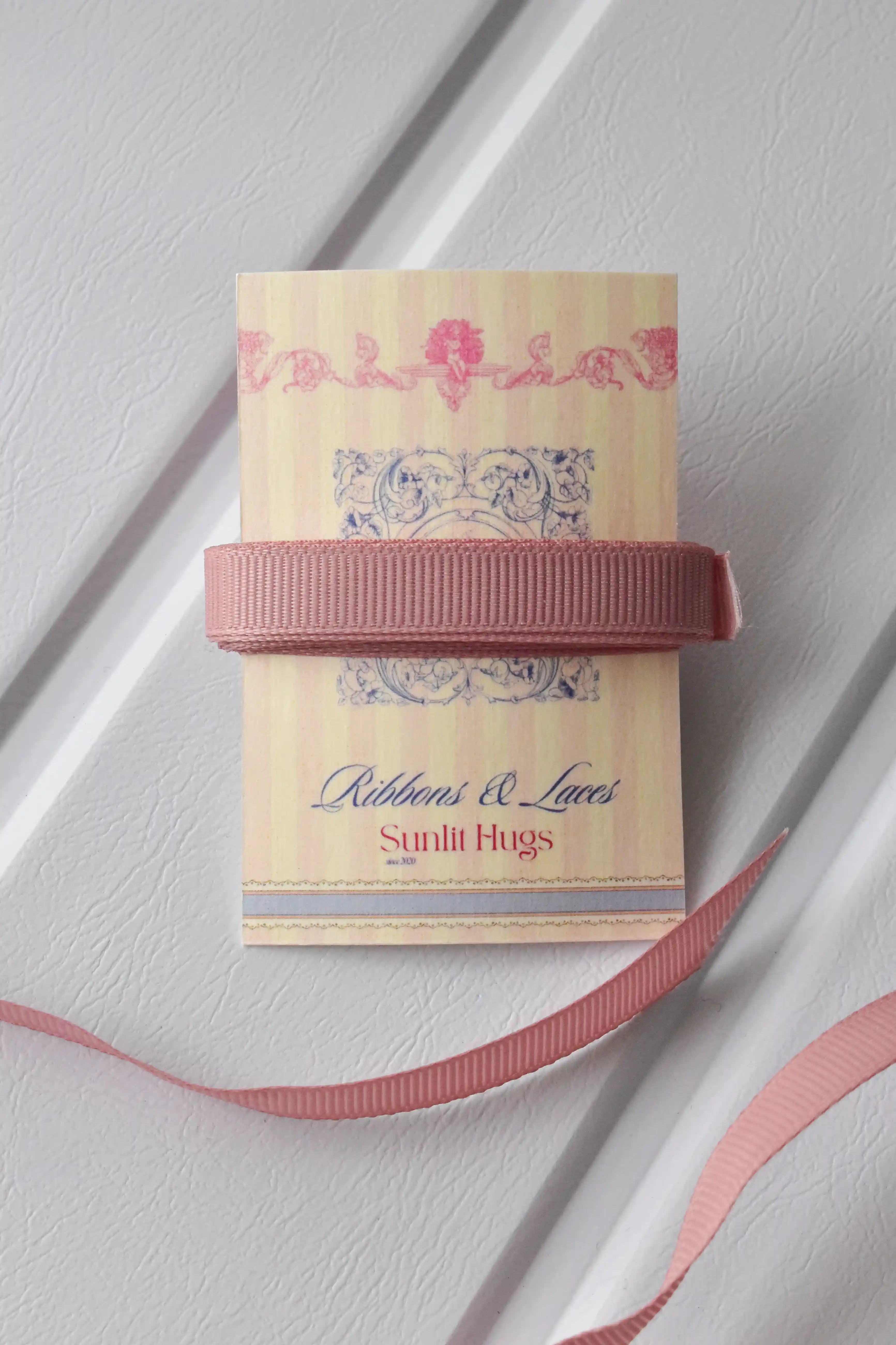 This image displays the Rustic Blush ribbon, showcasing its elegant rose gold colour and delicate texture. A long strand is loosely unravelled to highlight the full design, with a section wrapped around a "Sunlit Hugs" brand tag.