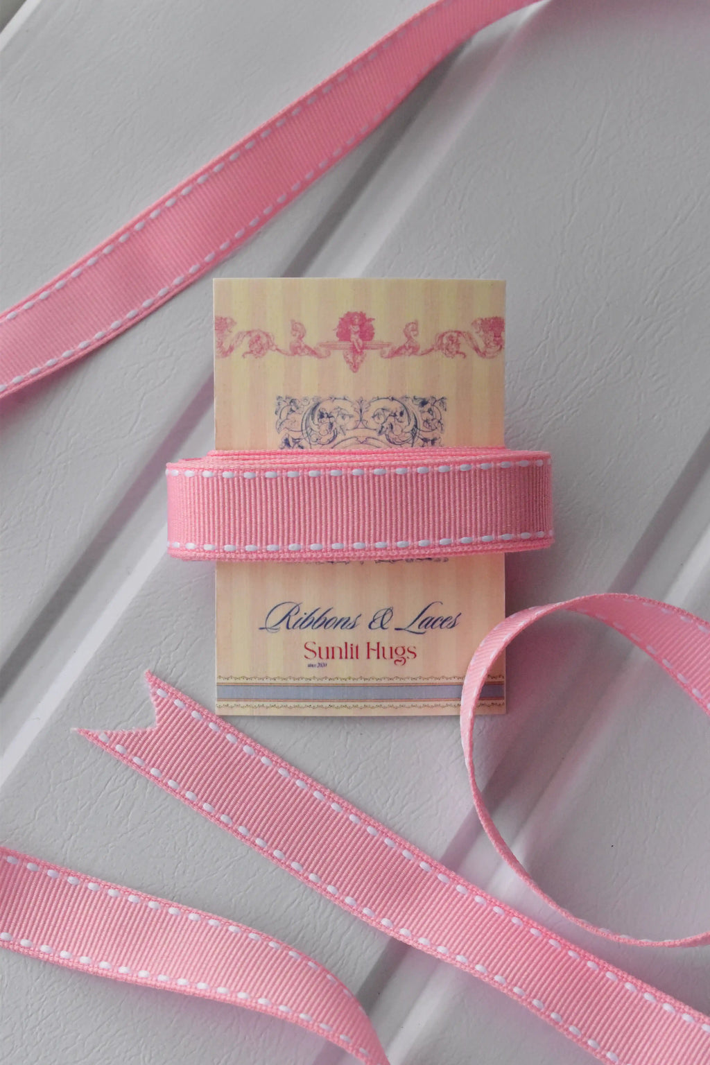 This image displays the Rose Taffy ribbon, showcasing its soft pink colour and delicate white dashed stitching. A long strand is loosely unravelled to highlight the full design, with a section wrapped around a "Sunlit Hugs" brand tag.