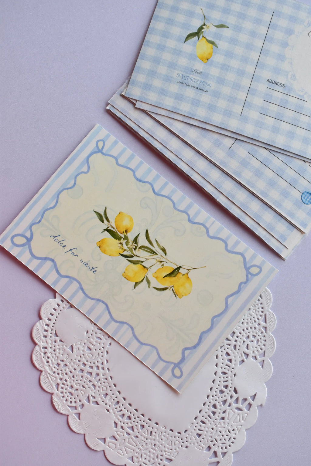 A set of postcards with the front 'Dolce Far Niente' lemon design laid flat on a white lace doily, and a stack of the blue gingham reverse sides visible behind it.