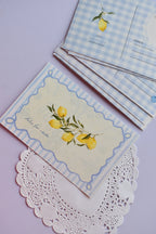A set of postcards with the front 'Dolce Far Niente' lemon design laid flat on a white lace doily, and a stack of the blue gingham reverse sides visible behind it.