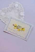 A close-up of the front of the postcard featuring bright, watercolor lemons on a branch, framed by a striped blue and scalloped border, with the phrase "dolce far niente" inscribed below.