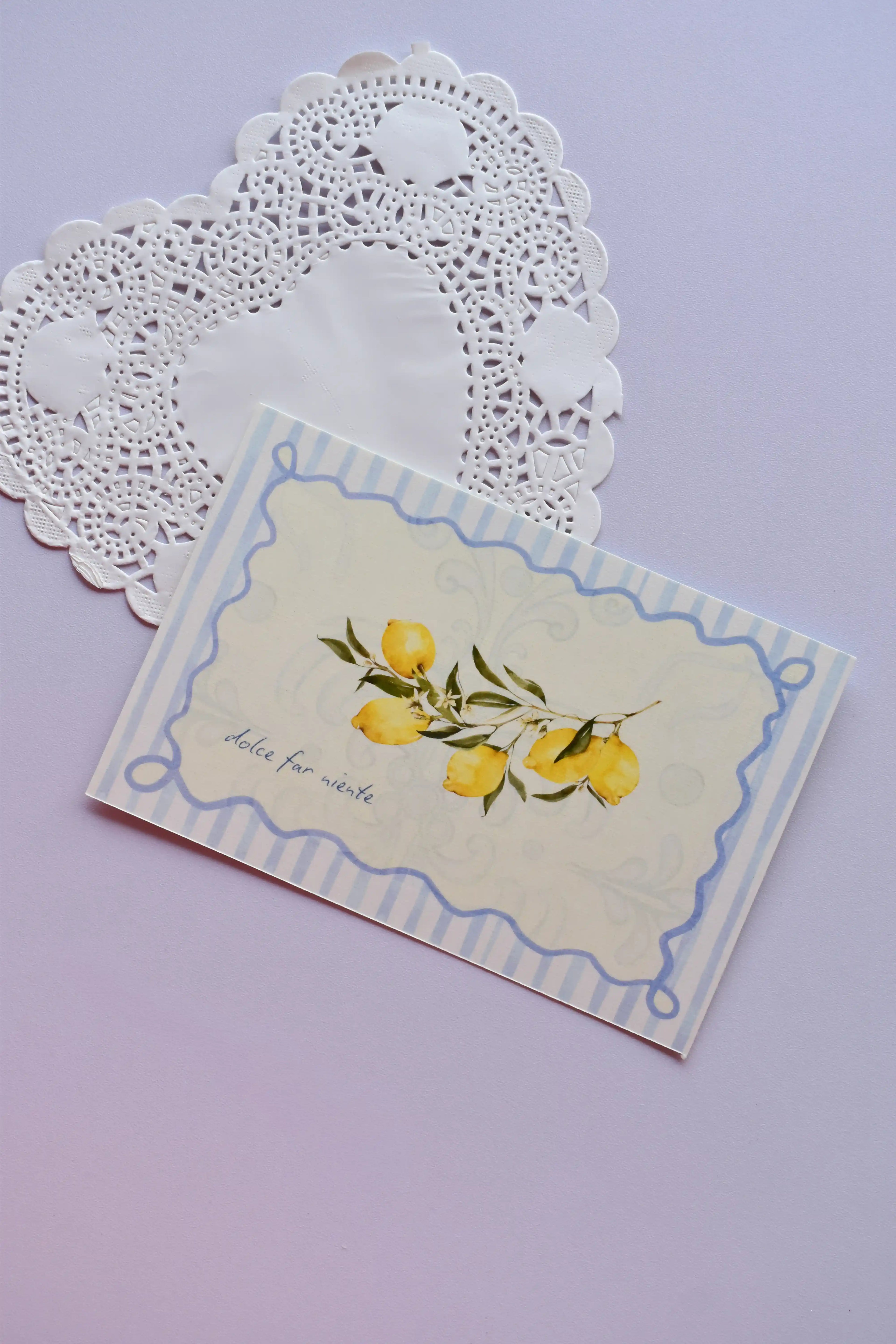 Close-up of a horizontal postcard featuring a watercolor illustration of yellow lemons on a branch. The image is framed by a decorative blue striped border and includes the text "dolce far niente." The postcard rests on a white heart-shaped paper doily.