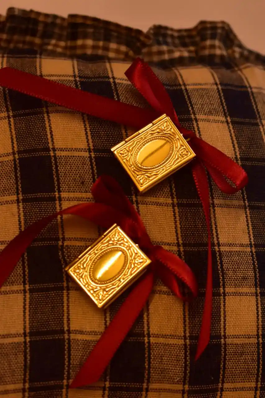 Pair of Golden Photo Book Pendant lockets tied with red satin ribbons, displayed on a vintage brown plaid pillow for a warm, nostalgic aesthetic.