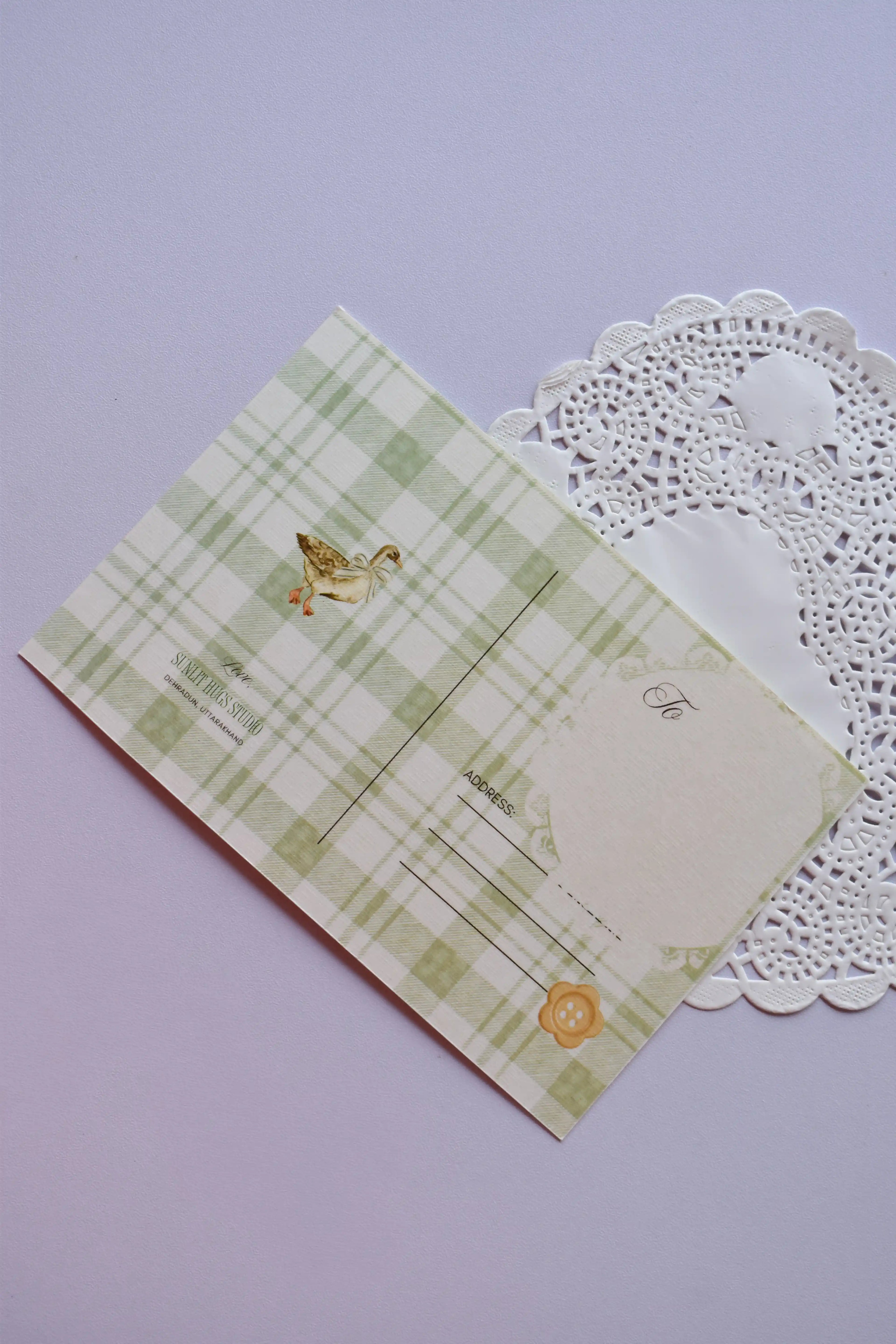 The back view of a postcard resting on a white paper doily. The design is a soft green and white wide plaid pattern, featuring a small illustration of a duck and a yellow button detail. It includes labeled lines for the address.