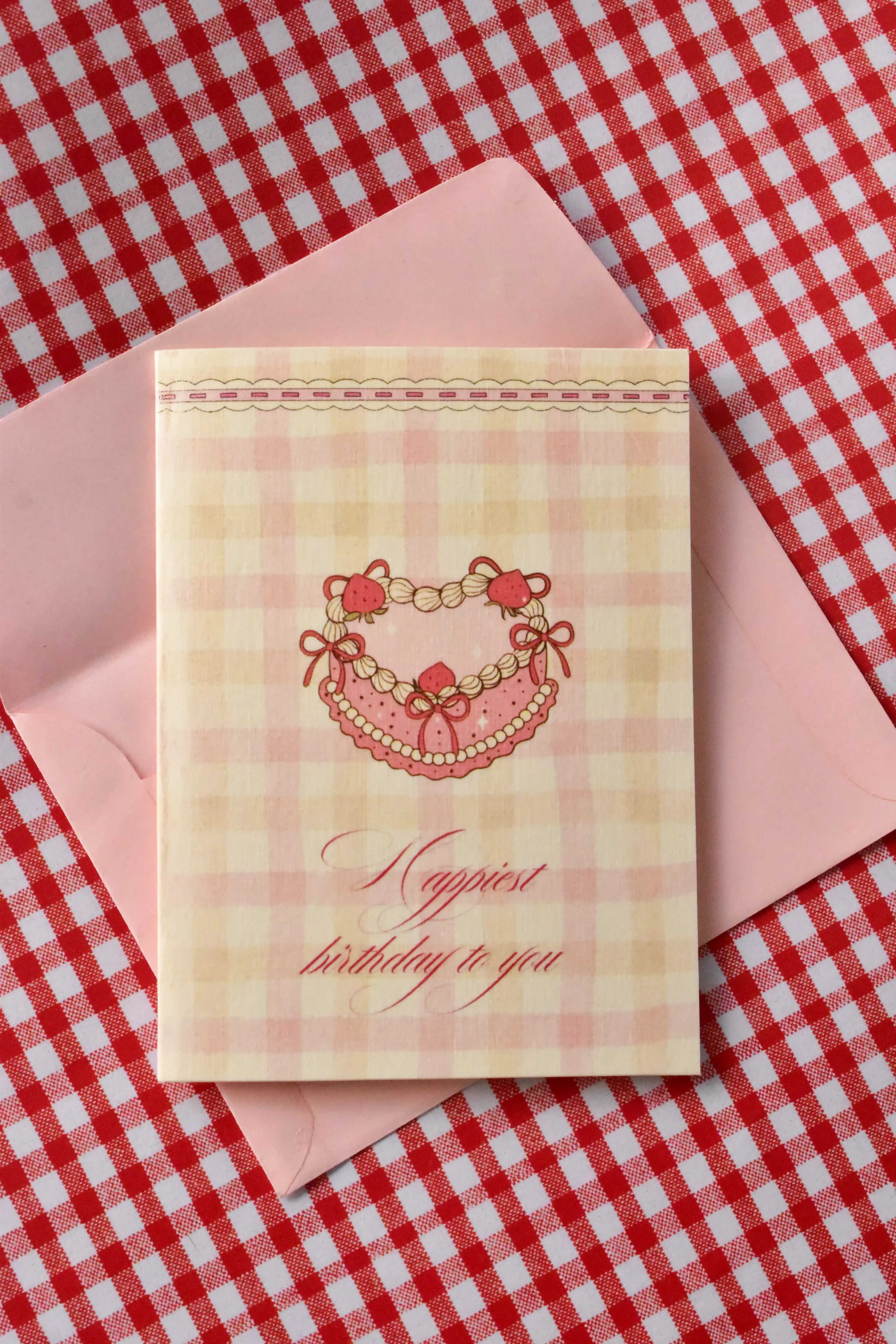 This image showcases the Happiest Birthday To You card, highlighting the sweet pink heart-shaped cake design and the cosy pink gingham pattern for pure birthday joy.