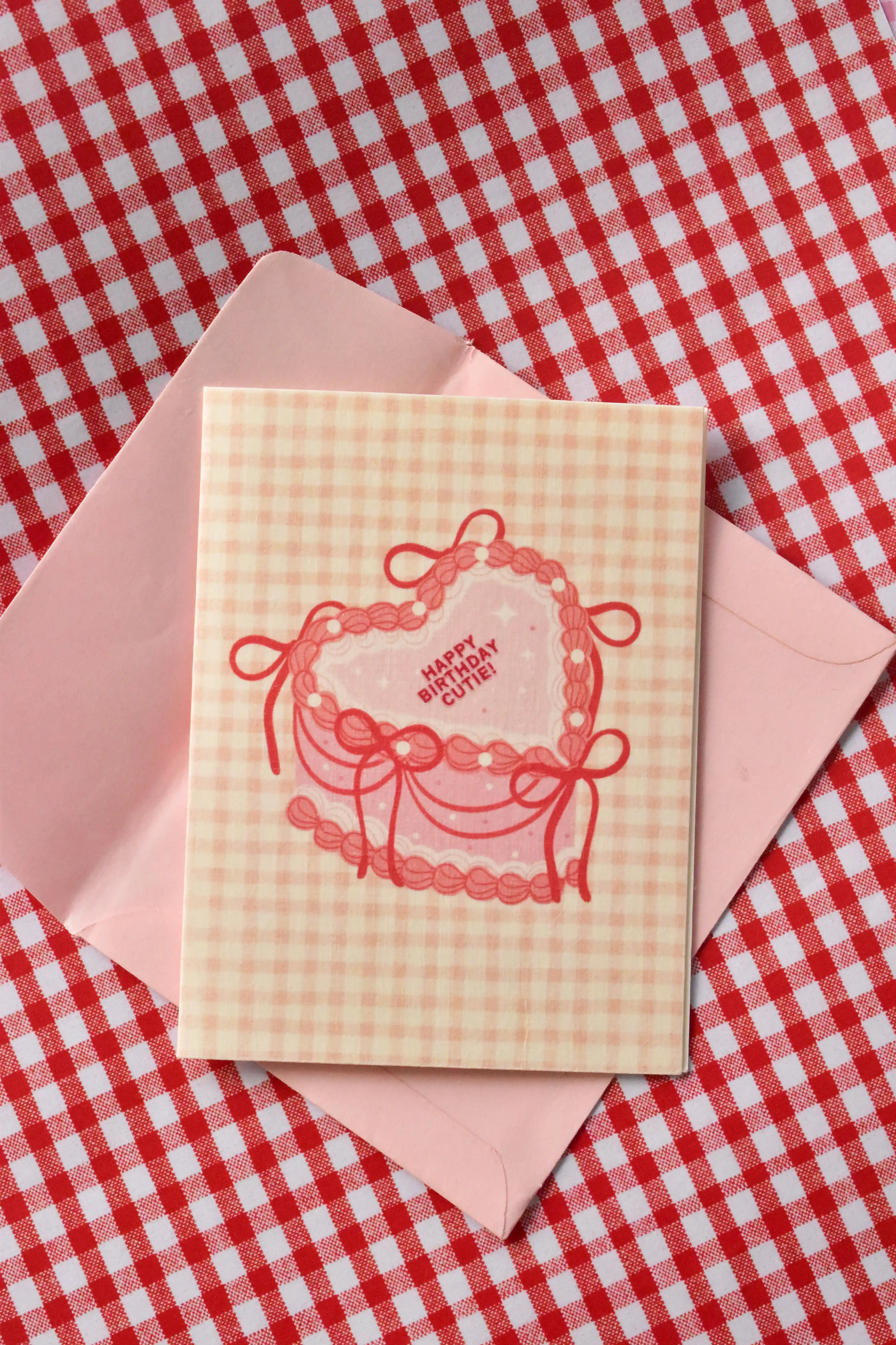 A close-up view highlighting the sweet pink heart-shaped cake design and the cosy pink gingham pattern of the 'Happy Birthday Cutie' card from the set.