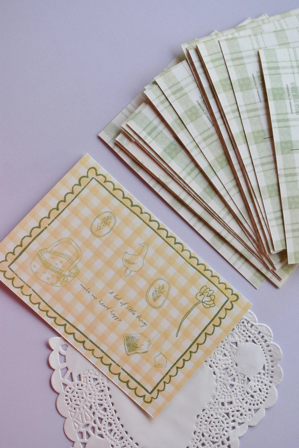 A clear view of the 'A lot of little things make my heart happy' postcard front, featuring yellow wide gingham and line-art sketches. A stack of the green plaid reverse sides, showing the duck and address lines, is arranged neatly above it.