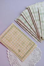 A clear view of the 'A lot of little things make my heart happy' postcard front, featuring yellow wide gingham and line-art sketches. A stack of the green plaid reverse sides, showing the duck and address lines, is arranged neatly above it.
