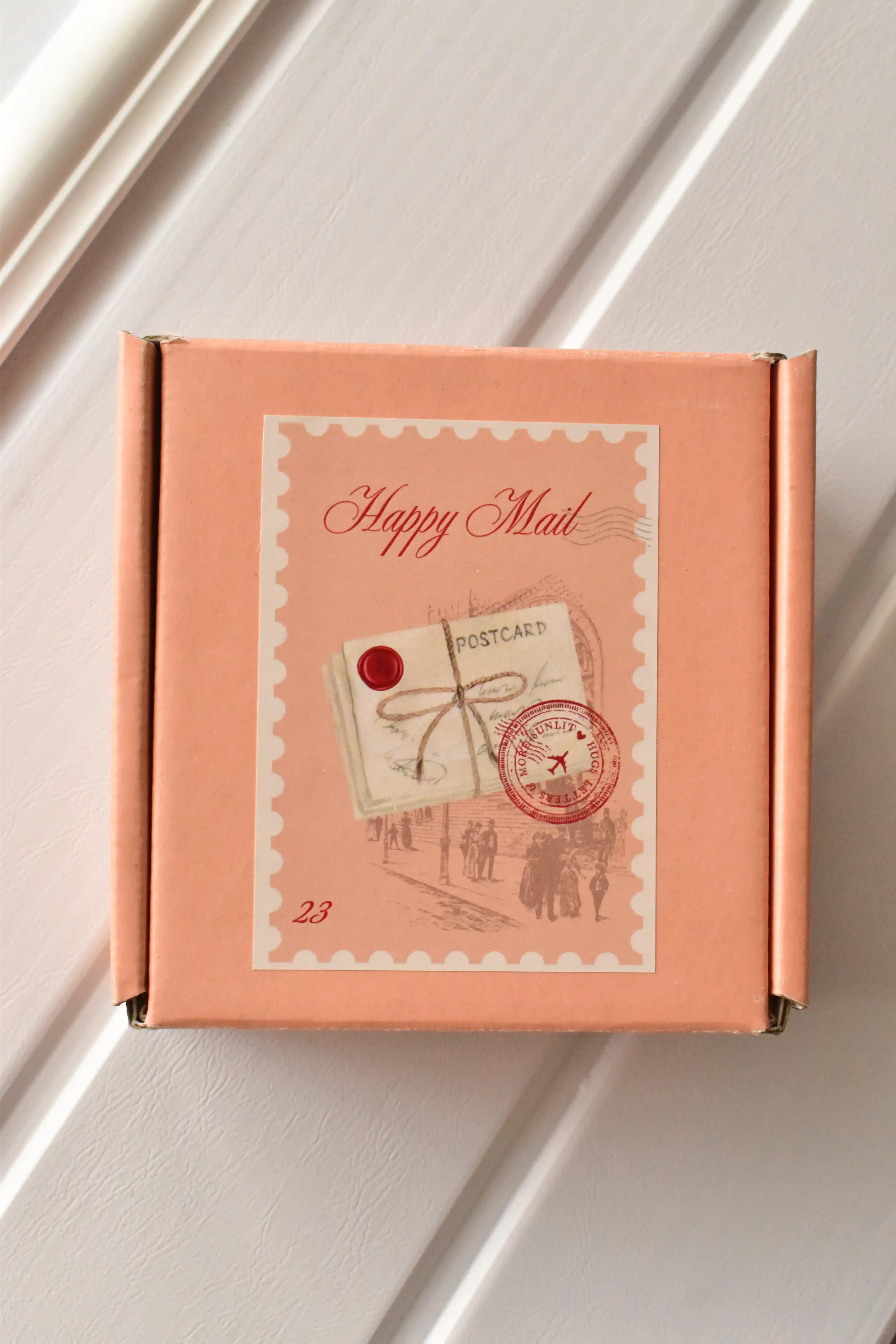 This top-down view showcases the charming 'Happy Mail' packaging, confirming that the bookmark will arrive in a delightful peach-coloured box with a vintage-style stamp.