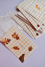 A horizontal postcard featuring a warm tan and cream wide gingham pattern. The front displays watercolor autumn leaves, an acorn, and the text: "In every leaf's shape, I find you." A stack of the reverse side of the postcards, showing the pie slice design, is visible behind it.