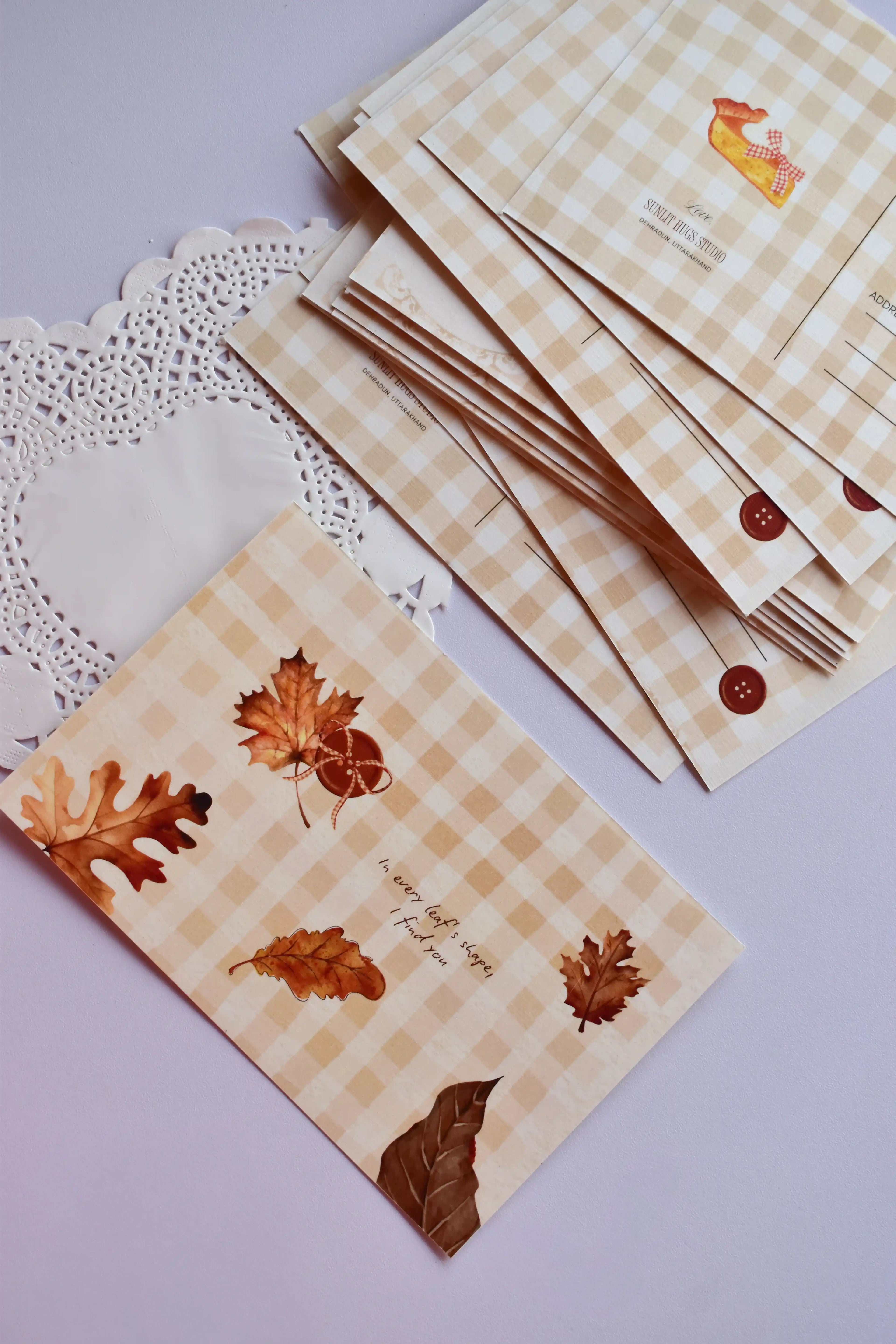 A horizontal postcard featuring a warm tan and cream wide gingham pattern. The front displays watercolor autumn leaves, an acorn, and the text: "In every leaf's shape, I find you." A stack of the reverse side of the postcards, showing the pie slice design, is visible behind it.