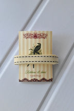 This detailed, head-on view of the Ivory Stitch ribbon highlights its soft cream colour and the contrasting brown dashed stitching. The ribbon is wrapped tidily around the "Sunlit Hugs" brand tag to show its precise design.