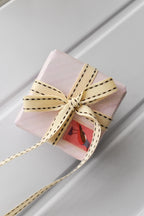 This image shows a beautifully wrapped gift box, featuring the Ivory Stitch ribbon tied in a bow. The cream and brown ribbon adds a sophisticated touch, with a vintage-style stamp placed underneath for a charming finish.