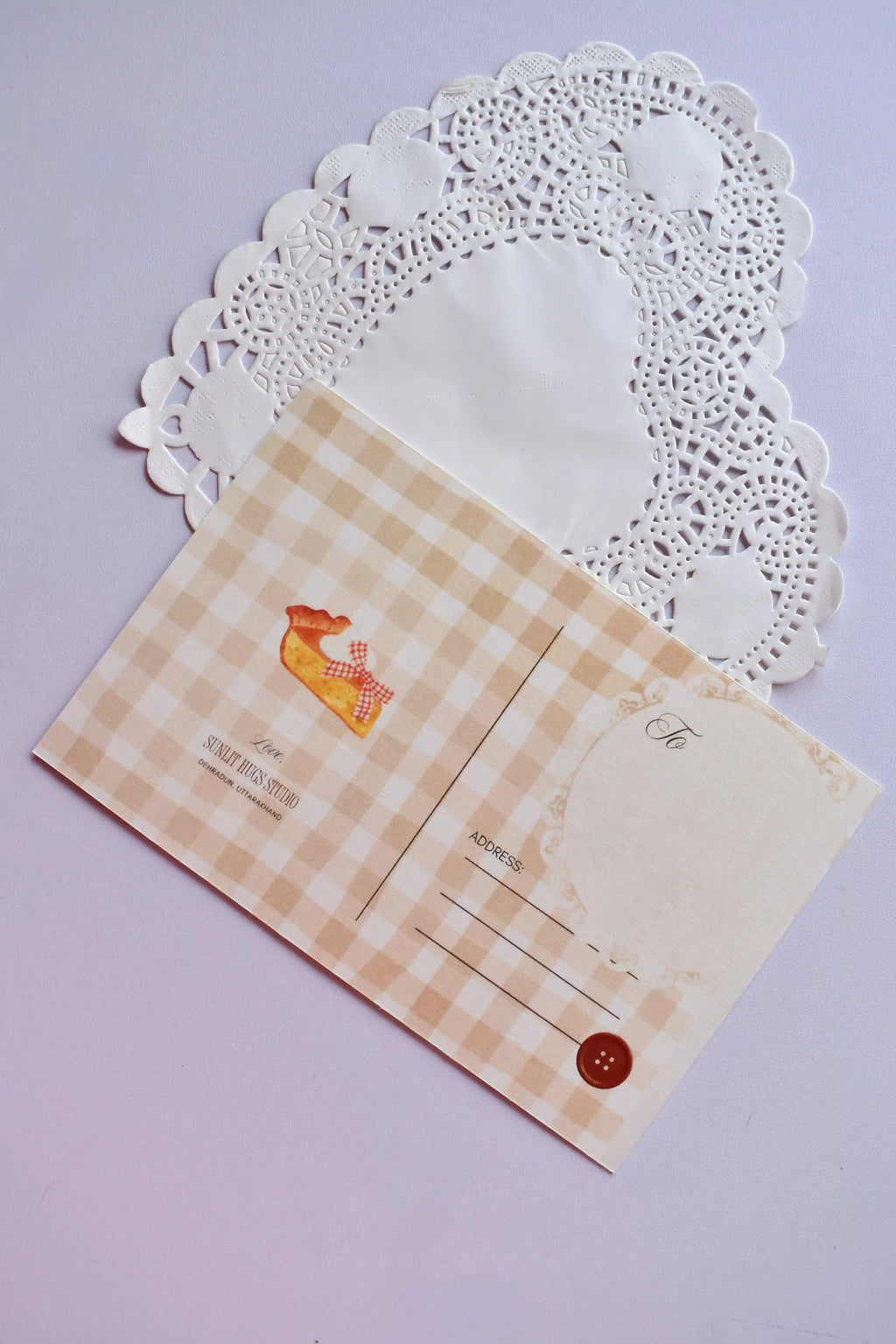 Close-up of the reverse side of a postcard on a white heart-shaped paper doily. The postcard has a tan and cream wide gingham pattern and features a small illustration of a slice of pie. It has sections for the address, postage, and a decorative brown button.