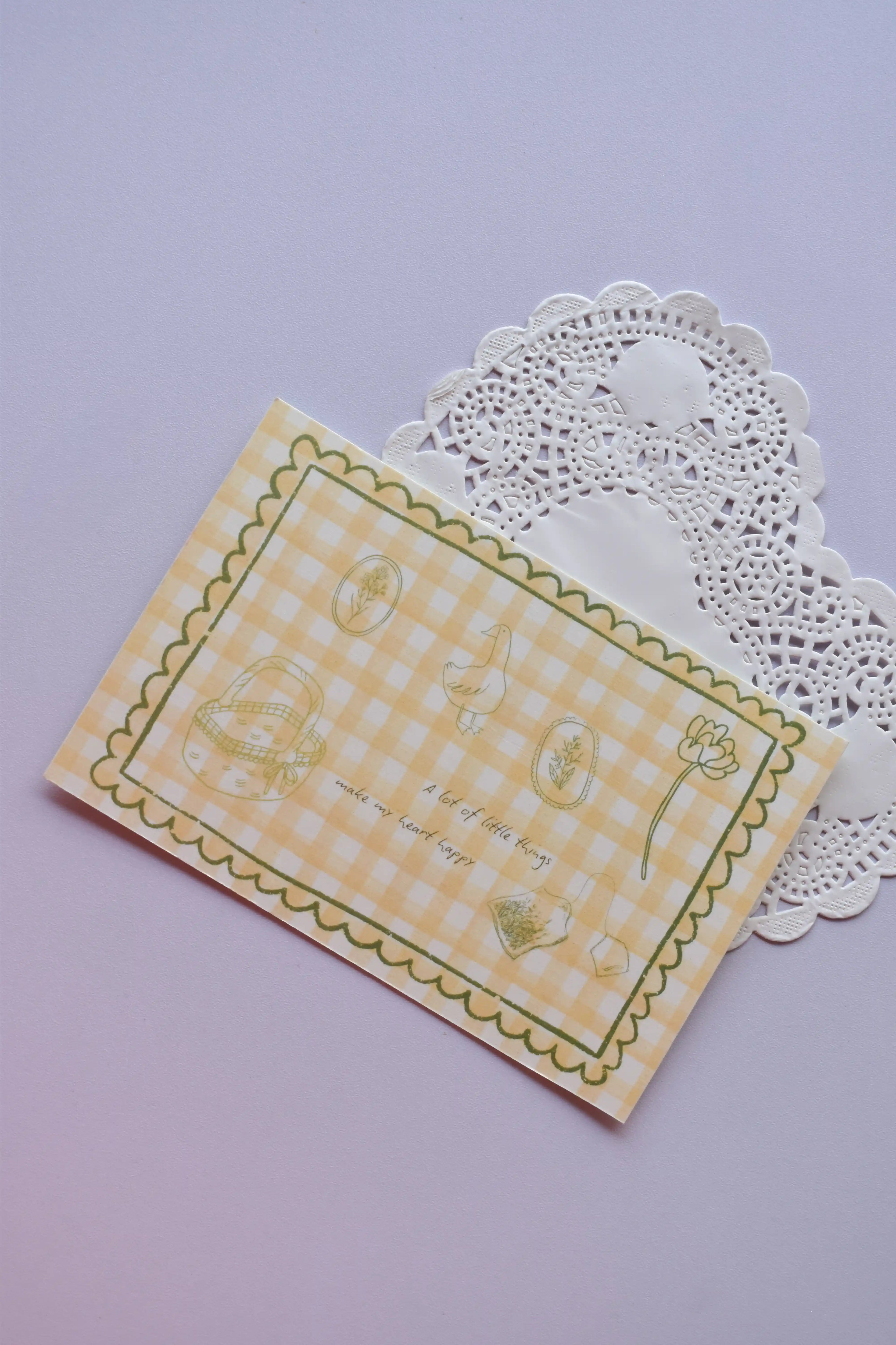 A horizontal postcard featuring a yellow and white wide gingham pattern with whimsical line-art sketches (a basket, a duck, and flowers) and the quote, "A lot of little things make my heart happy." A stack of the green gingham reverse side of the postcards is visible behind it.