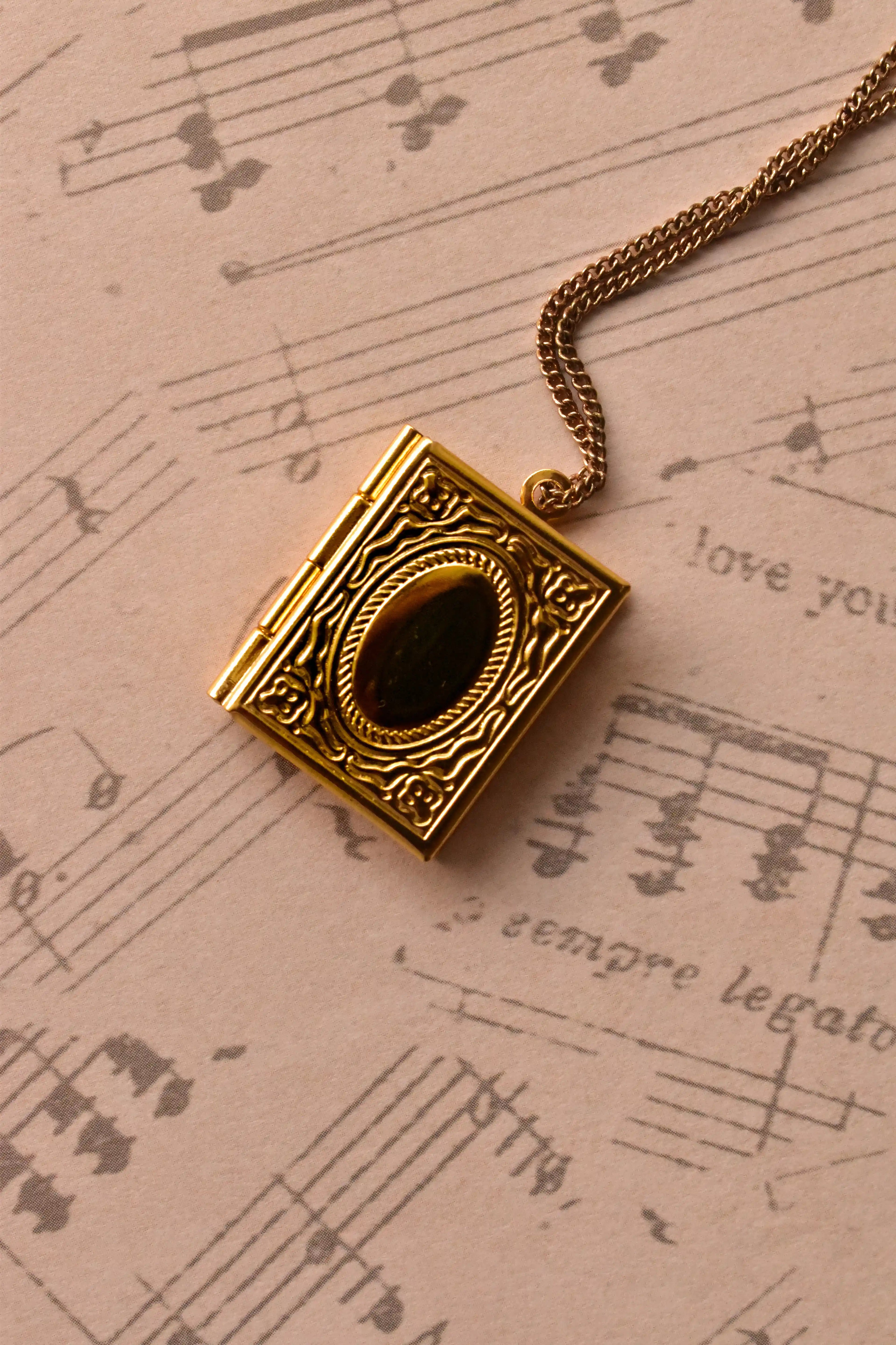 A close-up of the closed golden alloy locket, showcasing the intricate, book-style engraved design on the front, set against warm, nostalgic sheet music. (Note: Chain is for visual purposes only).