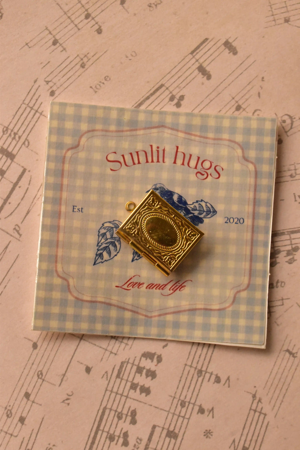 The golden alloy locket is presented on the Sunlit Hugs branded card, highlighting the detailed vintage design of the pendant itself.