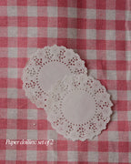 Set of two white paper doilies with a delicate lace border, resting on a pink and white checkered background.