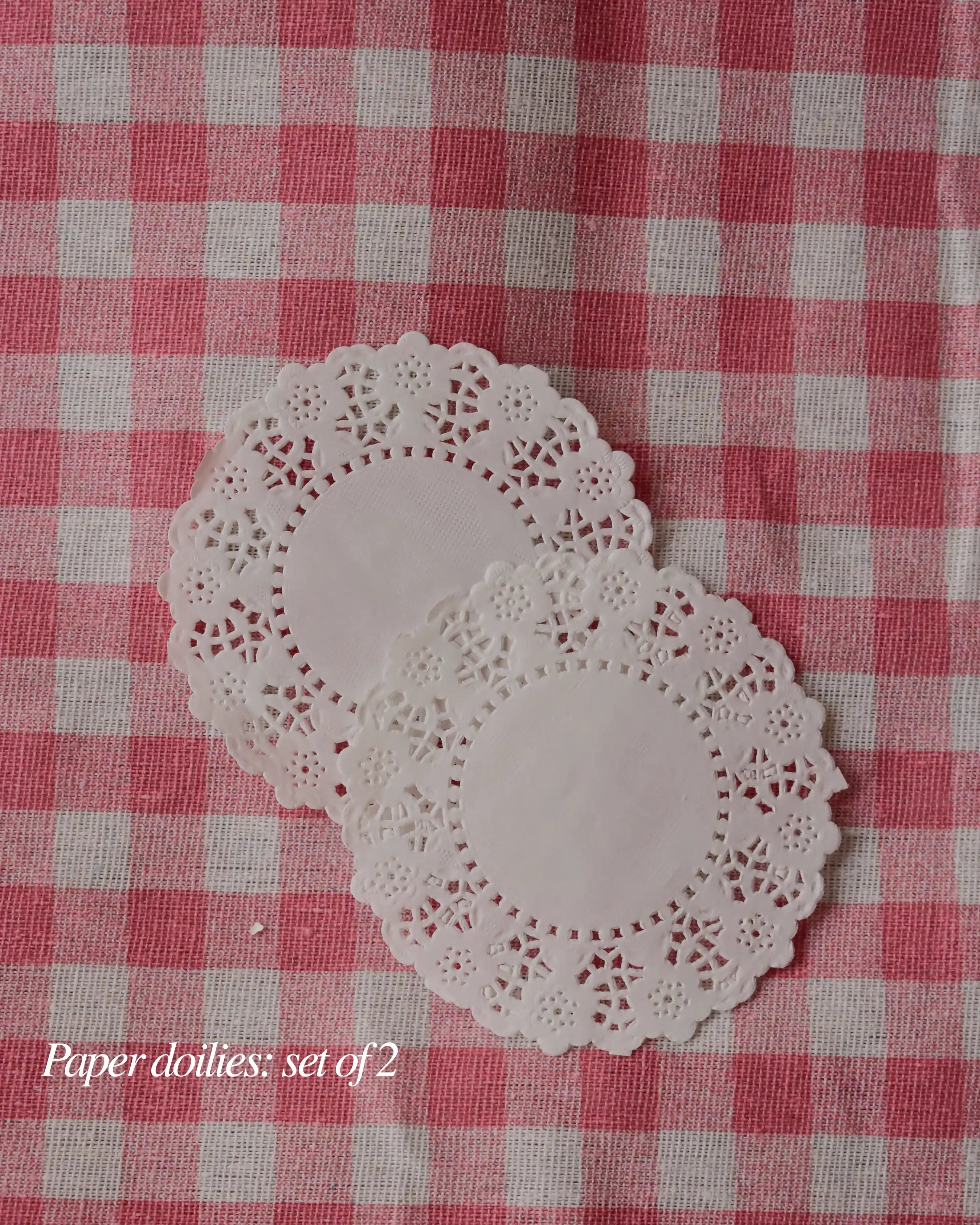 Set of two white paper doilies with a delicate lace border, resting on a pink and white checkered background.
