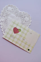 The back view of a postcard or a separate card design resting on a white heart-shaped paper doily. The design features a light green and white wide gingham pattern, with a decorative red checkered heart patch and a small green button detail. It includes lines for address and a spot for the postage stamp.