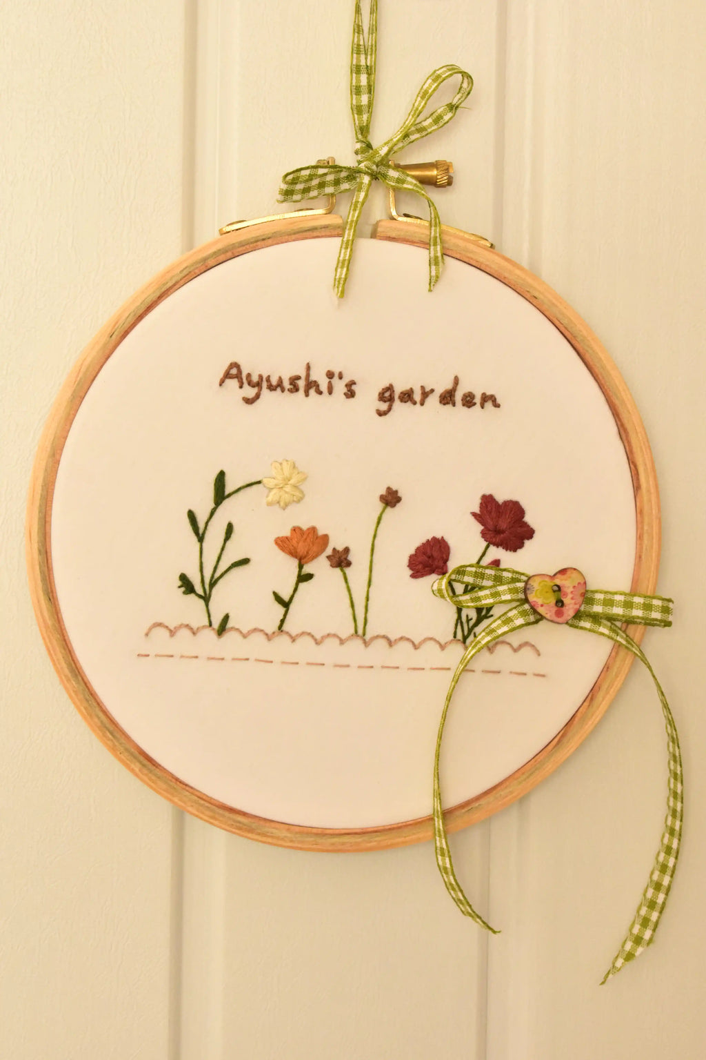 Custom 6-inch hand embroidered hoop reading "Ayushi's garden" with small flowers, hung on a white background.