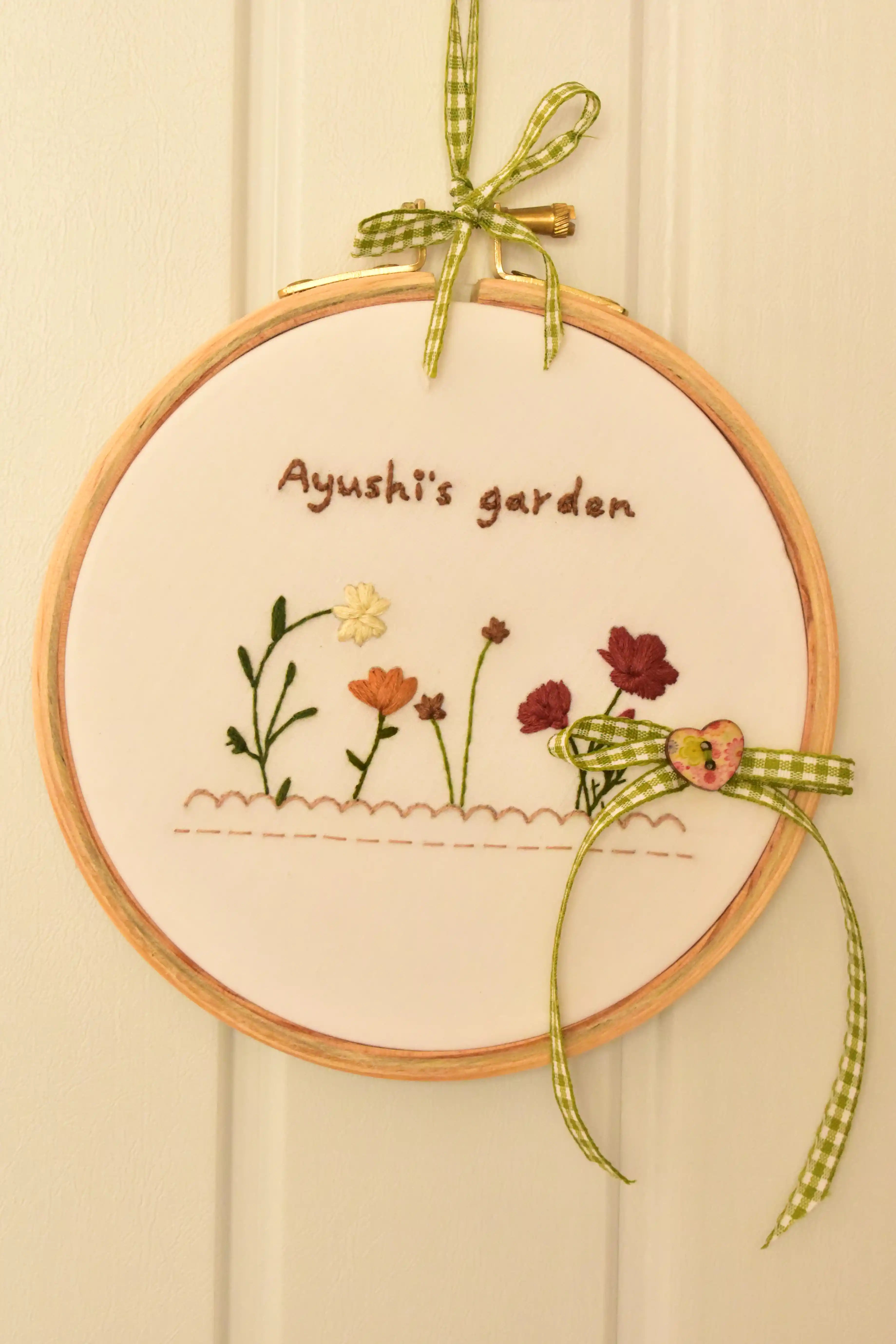 Custom 6-inch hand embroidered hoop reading "Ayushi's garden" with small flowers, hung on a white background.