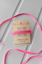 This image displays the Petal Satin ribbon, showcasing its vibrant pink colour and smooth texture. A long strand is loosely unravelled to highlight the full design, with a section wrapped around a "Sunlit Hugs" brand tag.