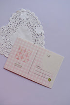 The back view of a postcard resting on a white paper doily. The design is a soft pink and white wide gingham pattern, with a central square featuring a pink floral bell pattern. It includes labeled fields for address and postage.