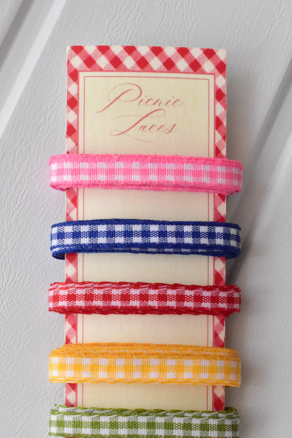 Vertical stack of five narrow gingham ribbons in pink, blue, red, yellow, and green on a "Picnic Laces" display card.